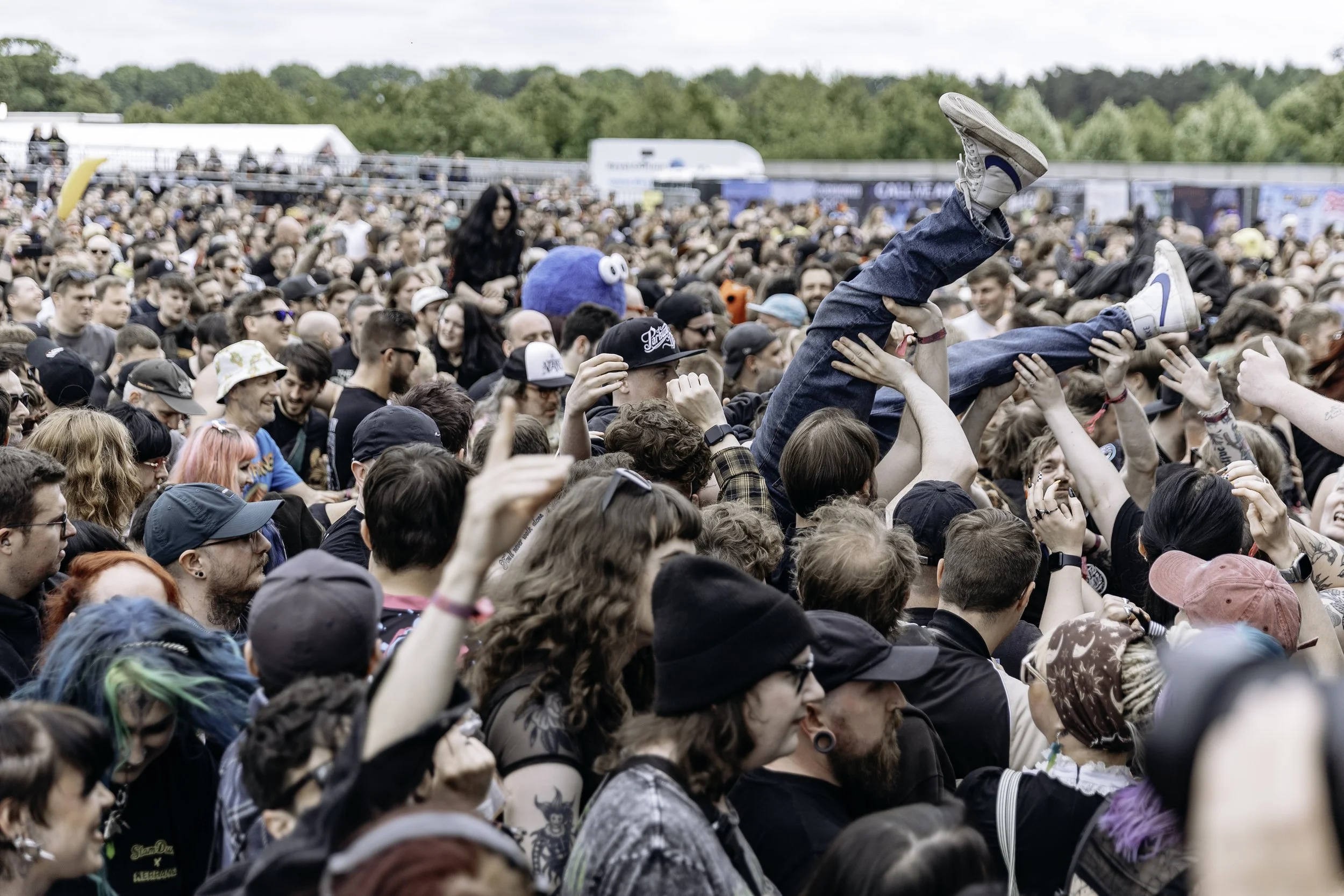 Crowd at outdoor music festival, with a person being lifted above the crowd and wearing a blue hat with white eyes.
