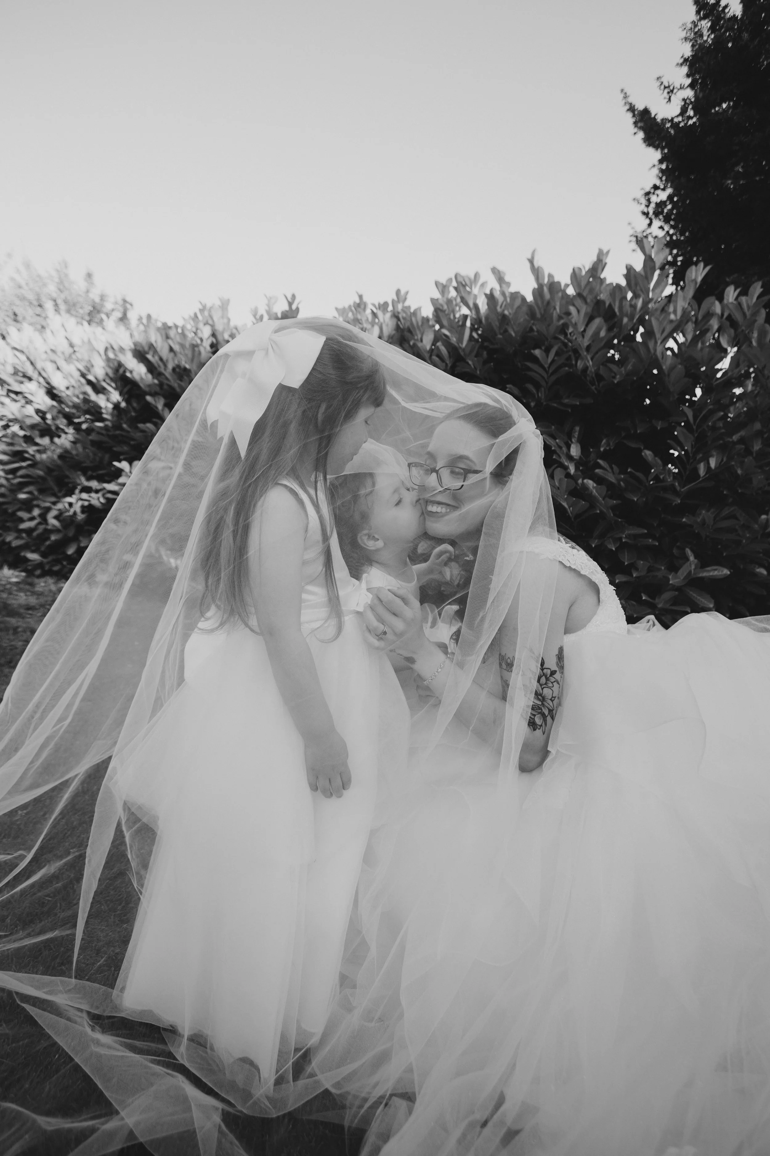 A woman in a wedding dress, two young girls, and a girl princess dress standing outside behind bushes, all under a sheer veil.