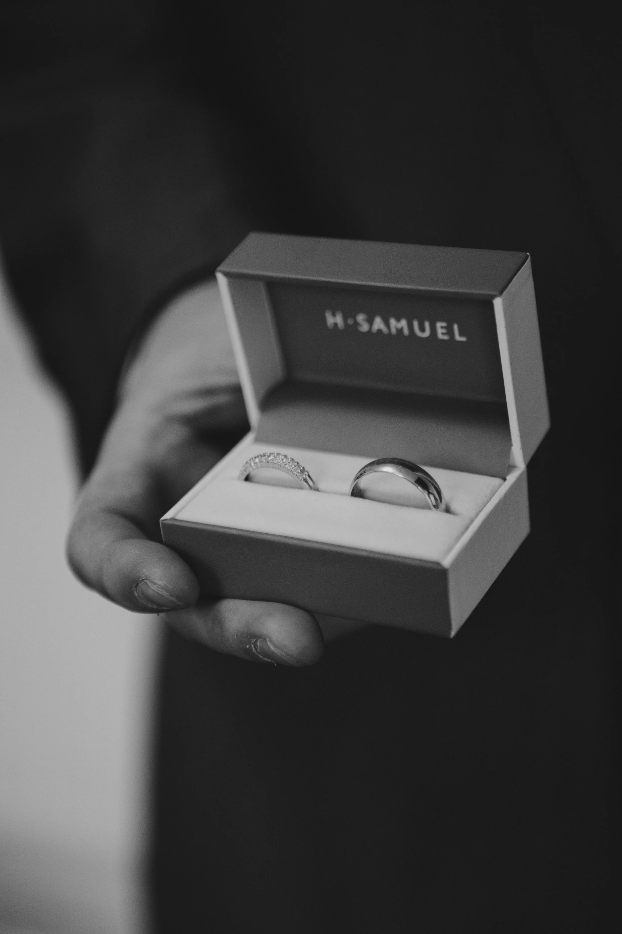 A hand holding an open jewelry box with two wedding bands, one with diamonds, inside. The box has the brand name 'H-SAMUEL' inside the lid.