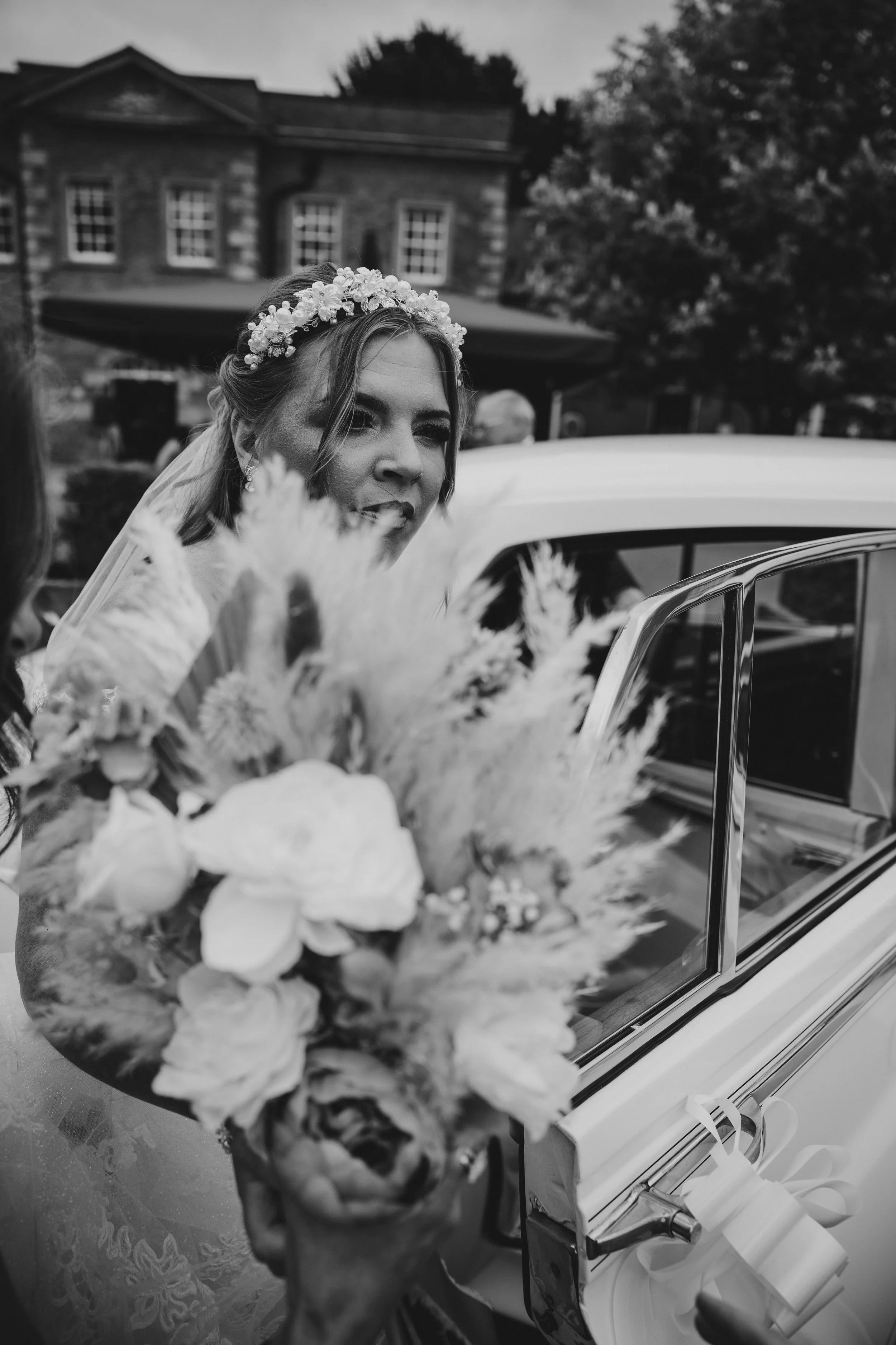 Black and white photo of a smiling bride wearing a floral crown and veil, holding a bouquet of flowers, as she stands next to a decorated car.