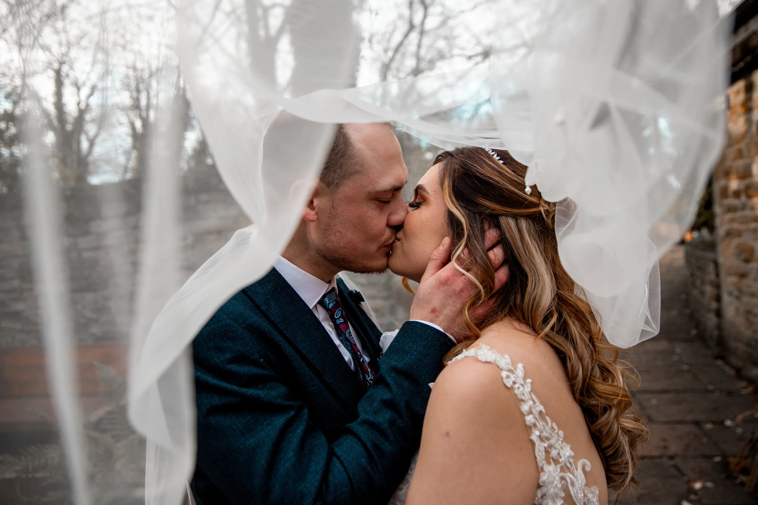 A bride and groom kiss surrounded by a flowing veil outdoors on a cloudy day.