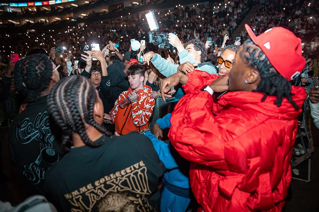 Group of people at a crowded event, taking photos and videos with their phones, illuminated by camera flashes.