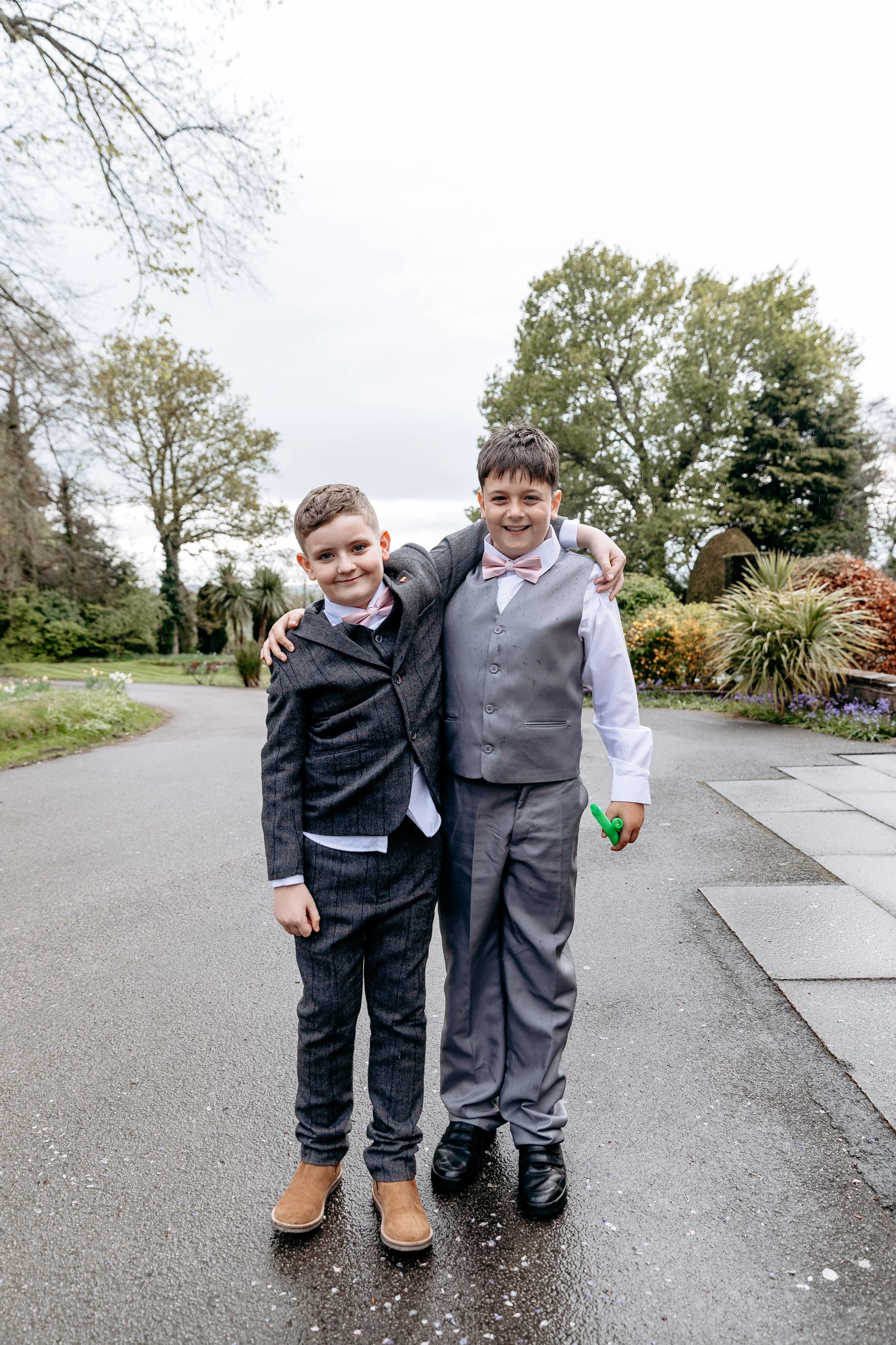 Two young boys dressed in formal suits smiling and standing close together outdoors in a garden or park, with trees and shrubs in the background.