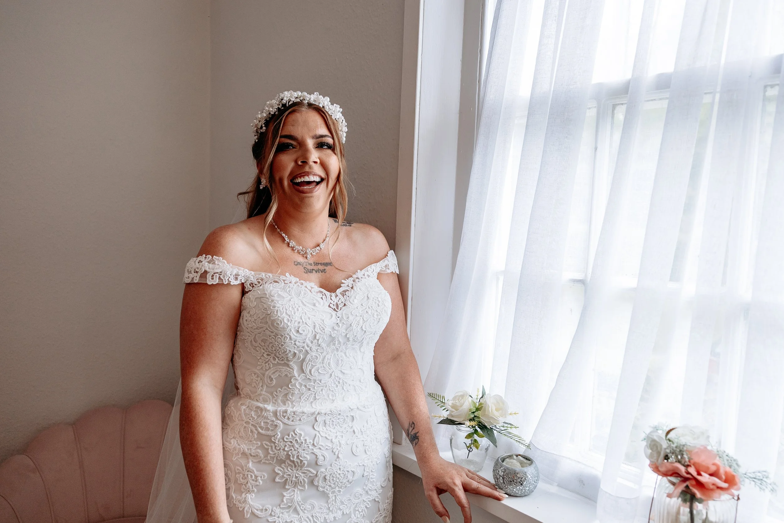 A smiling woman in a white lace wedding dress standing next to a window with sheer curtains, holding a small flower arrangement, with flowers and candles on the windowsill.