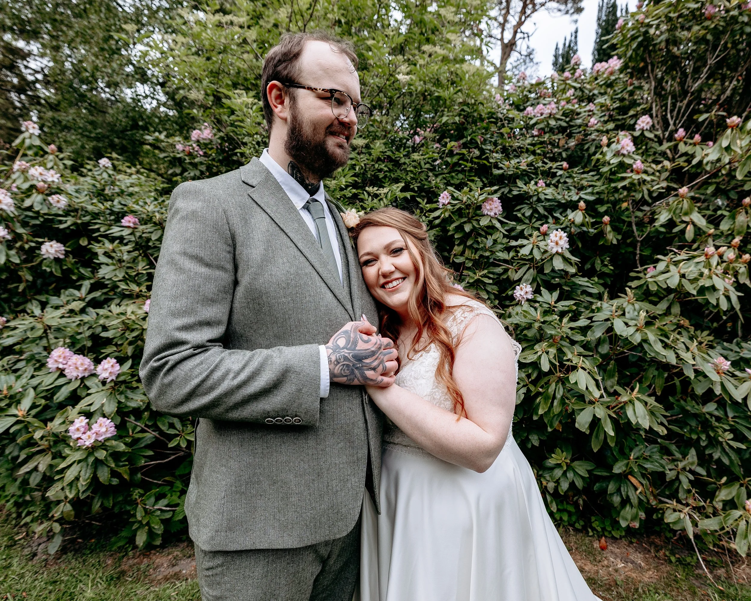 A happy bride and groom holding hands and smiling outdoors in front of flowering bushes.