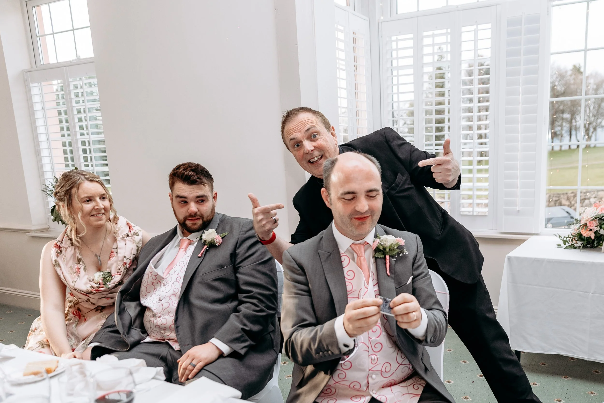 Group of five people at a wedding reception, three men and two women, sitting at a table, with one man standing behind them. All are wearing formal attire with floral boutonnieres or corsages. The man standing is pointing at one of the seated men, wh
