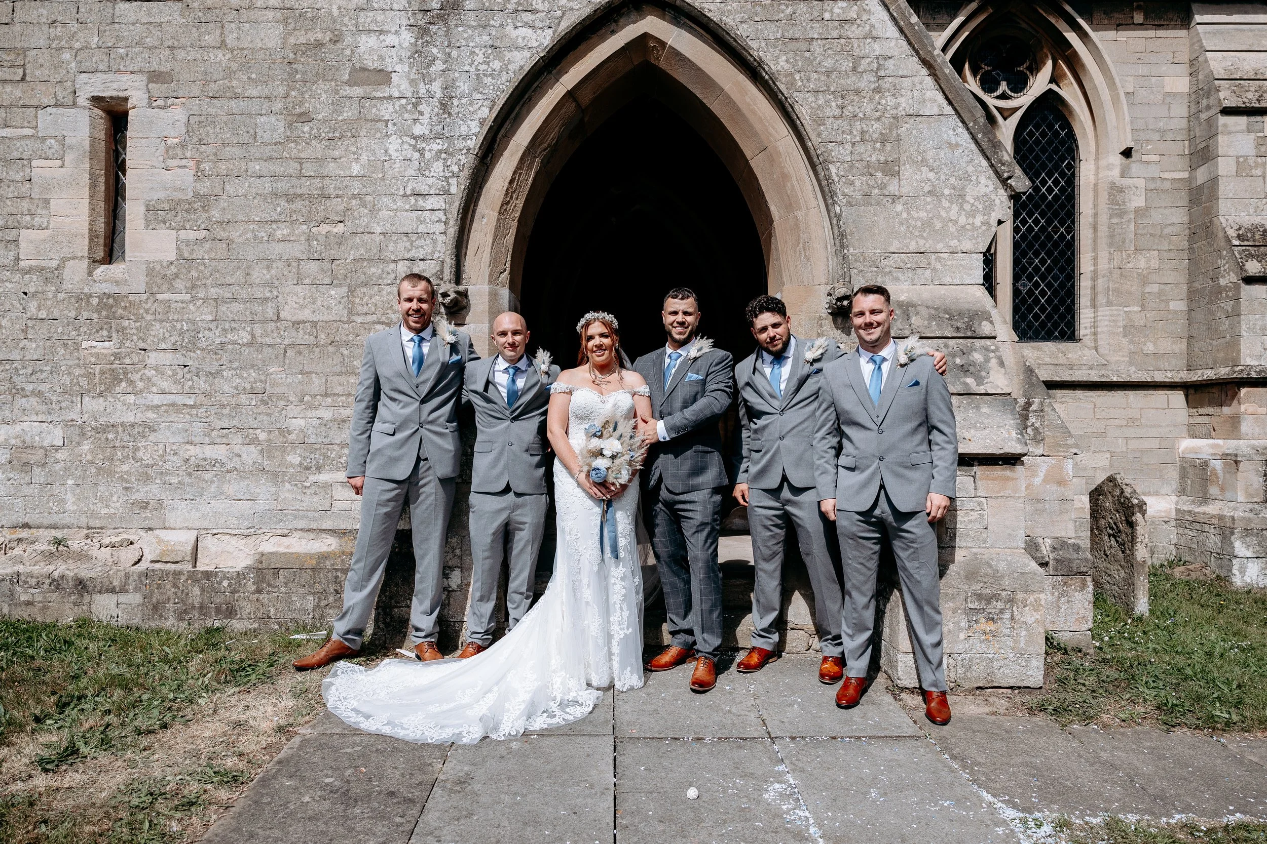 A bride and groom with five groomsmen in gray suits pose in front of a stone church during a wedding celebration.