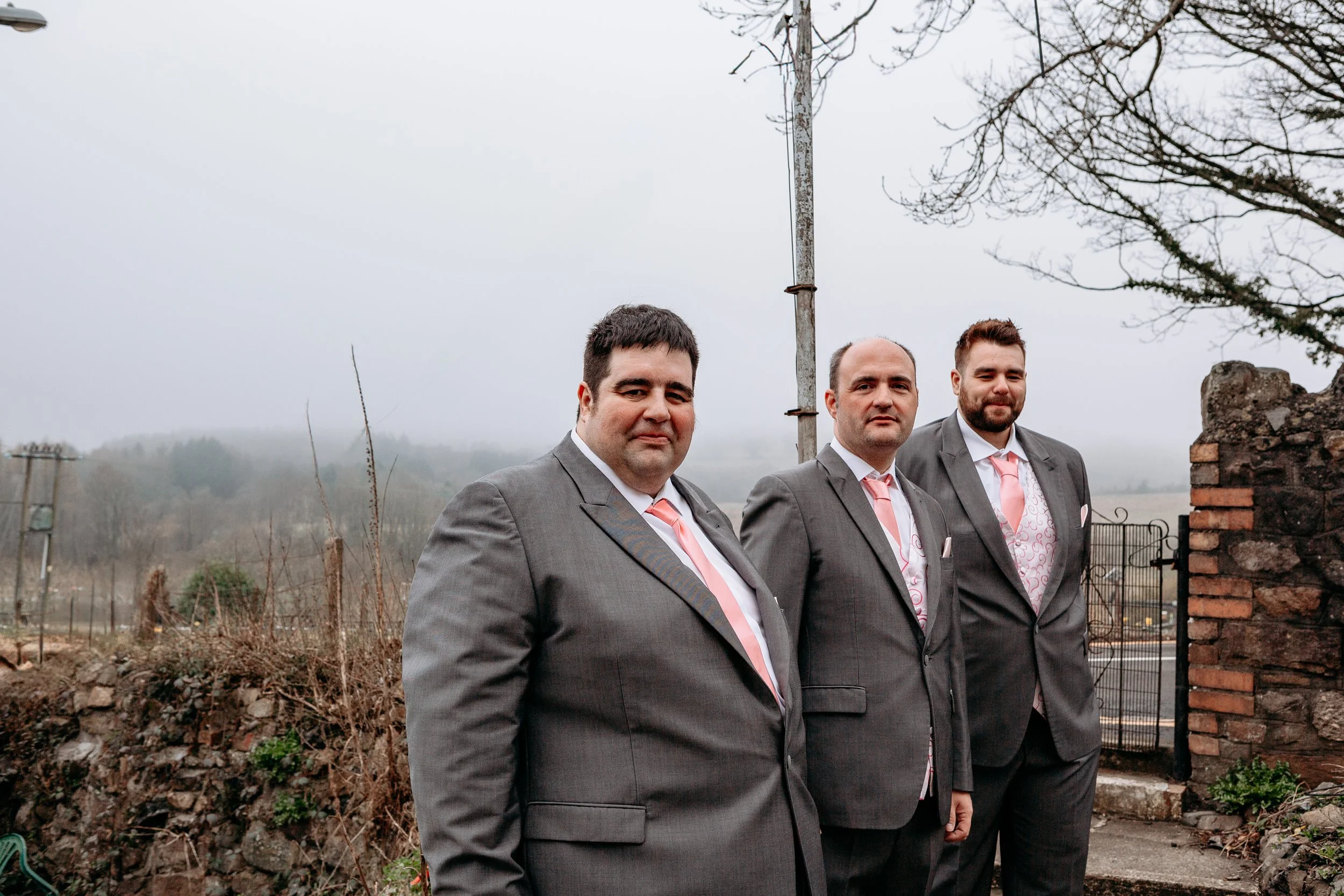 Three men dressed in gray suits with pink ties and pocket squares standing outdoors on a cloudy day near a brick wall and gate.
