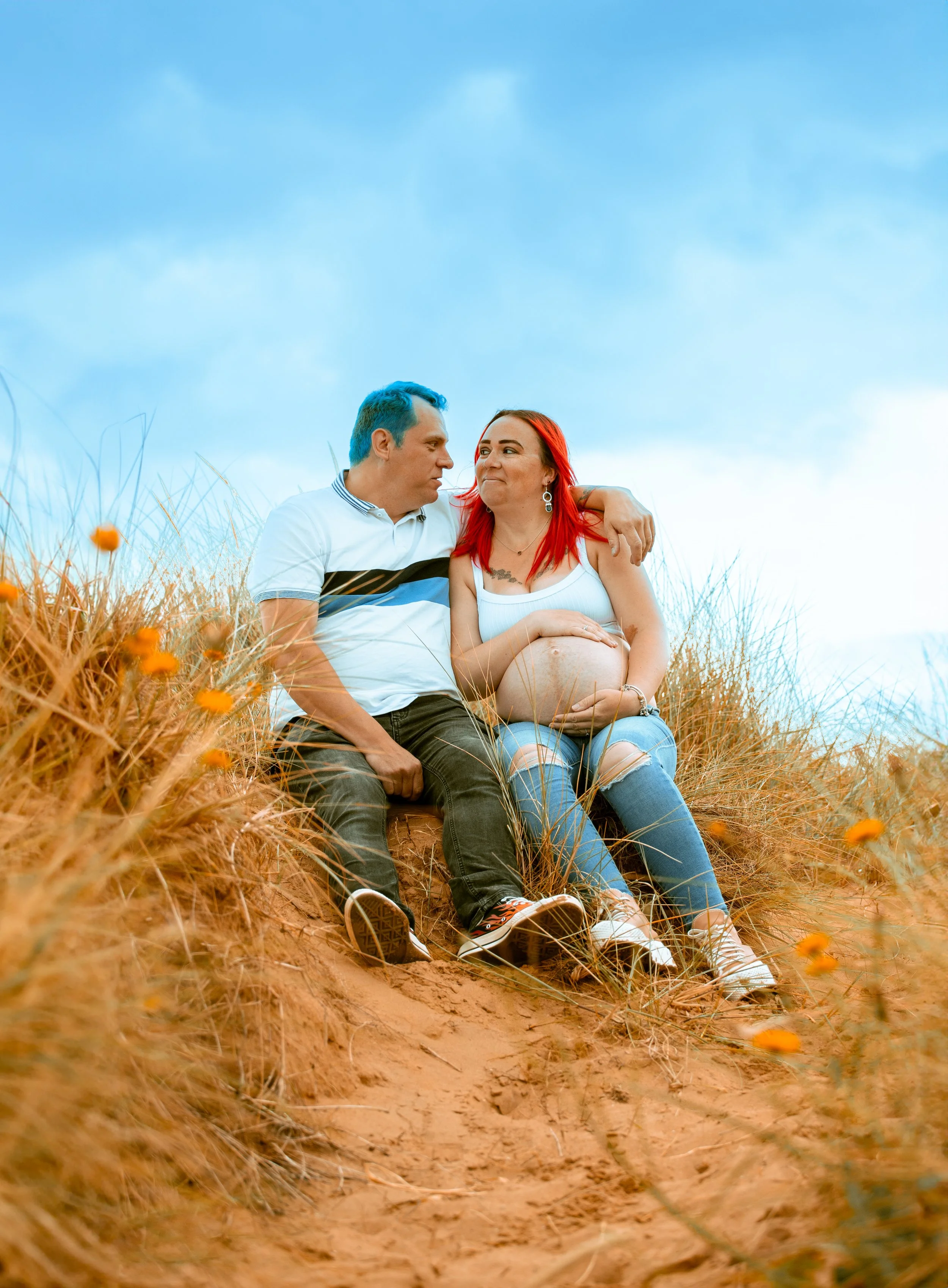 A couple sitting on a sandy hill, with the woman showing her pregnant belly, surrounded by dry grass and small orange flowers, under a blue sky.