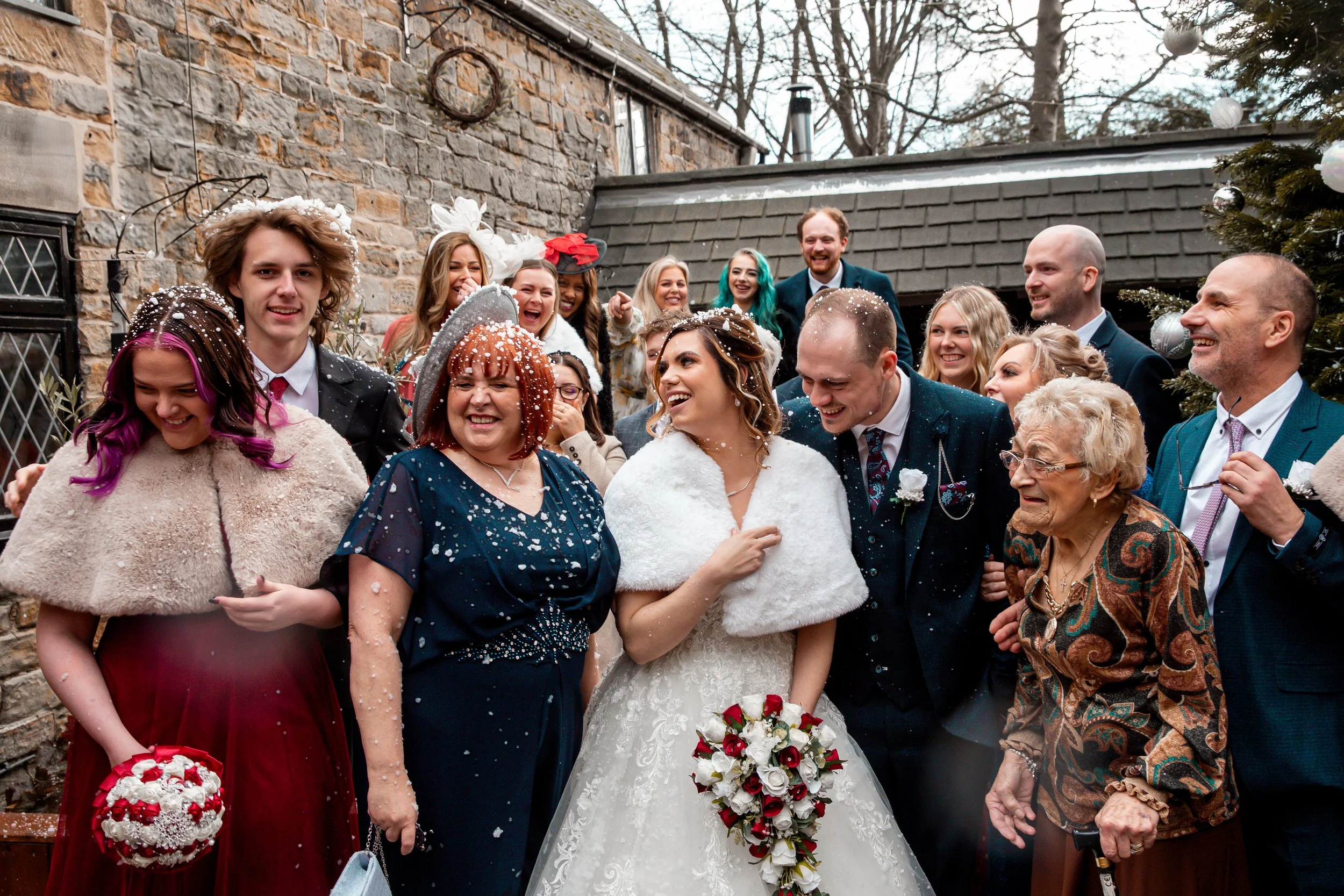 Group of people celebrating at a wedding, with a bride holding a bouquet, surrounded by family and friends, some throwing confetti, outdoor setting with stone wall and Christmas tree.