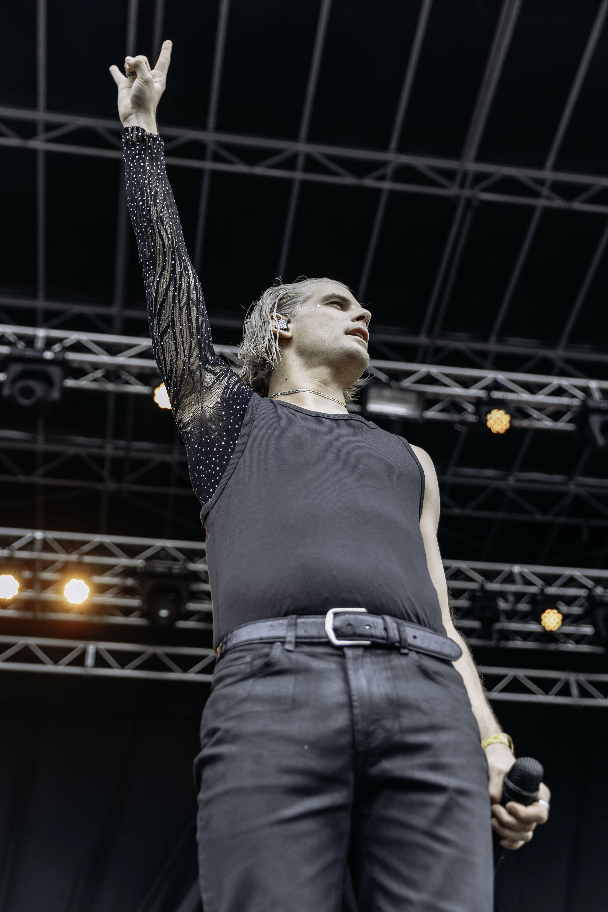 A male performer with platinum blonde hair on a stage, wearing a black sleeveless top with one sleeve made of sheer fabric with glittery patterns, holding a microphone in his right hand, looking upward, with stage lights and metal scaffolding in the 
