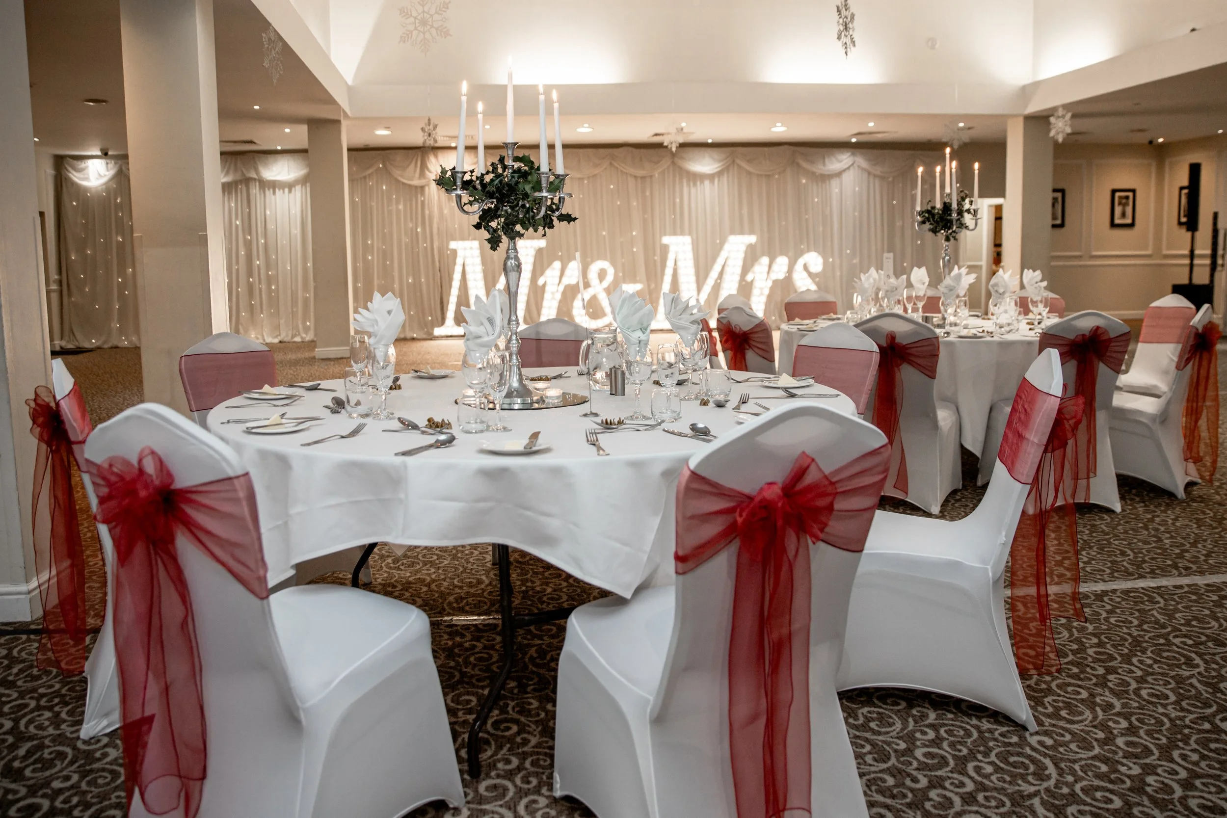 Elegant wedding reception setup with round tables covered in white tablecloths, surrounded by chairs wrapped in white covers with red ribbons, and centerpieces featuring tall silver candleholders with white candles. The background includes illuminate