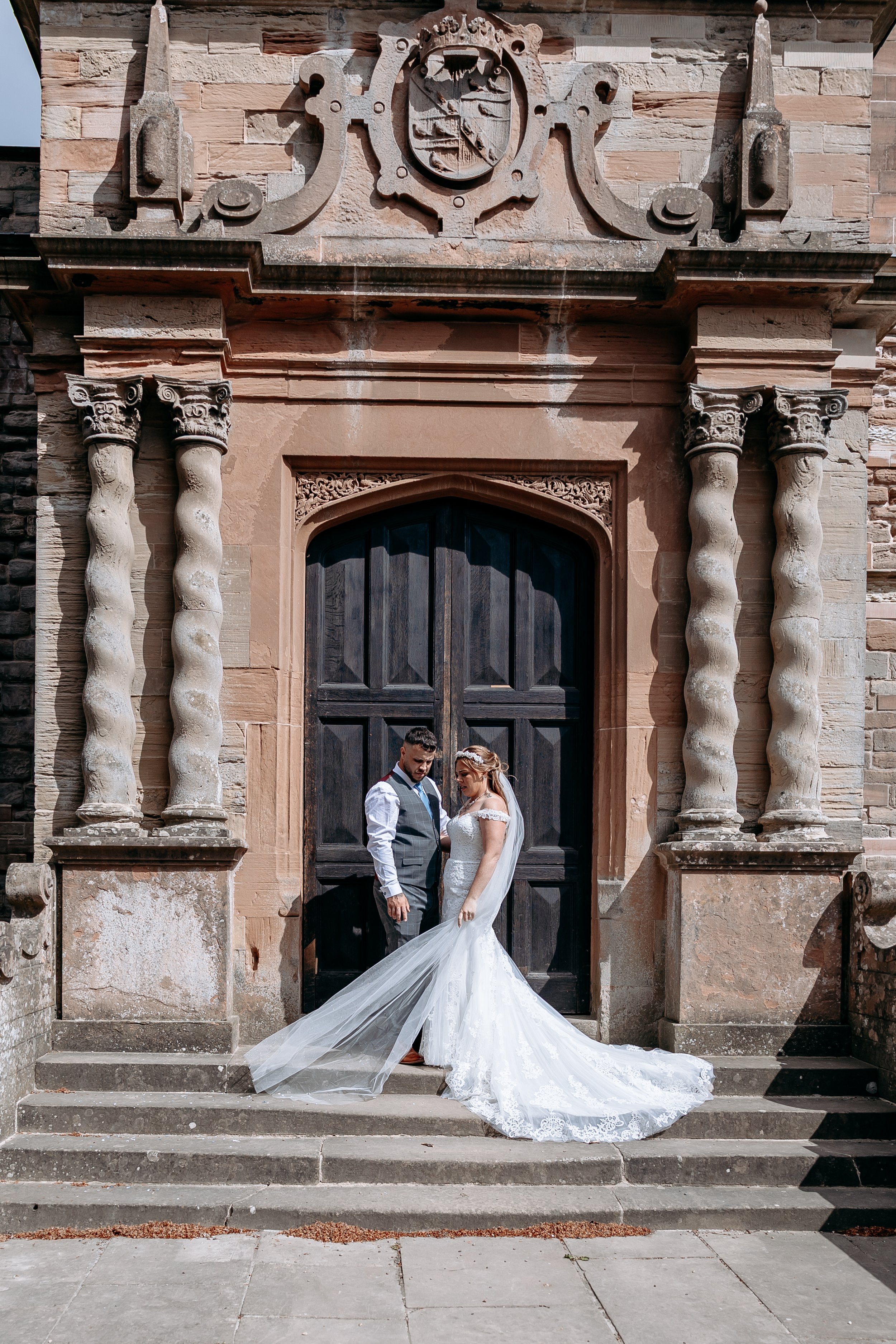 Bride and groom standing on stone steps in front of large wooden door with stone columns and ornate stone crest above, during their wedding.