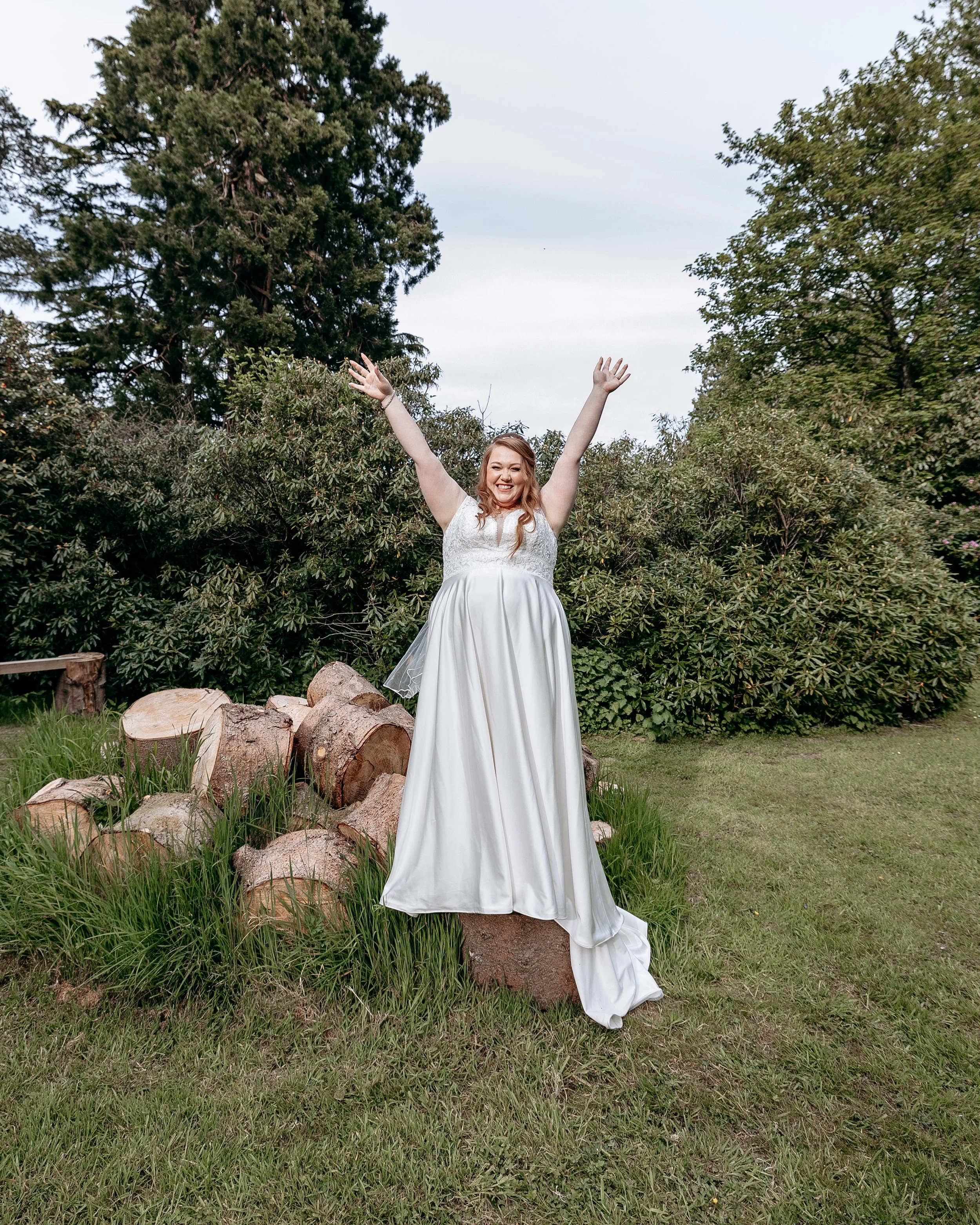 A happy woman in a white wedding dress standing outdoors with arms raised, surrounded by greenery and chopped logs.