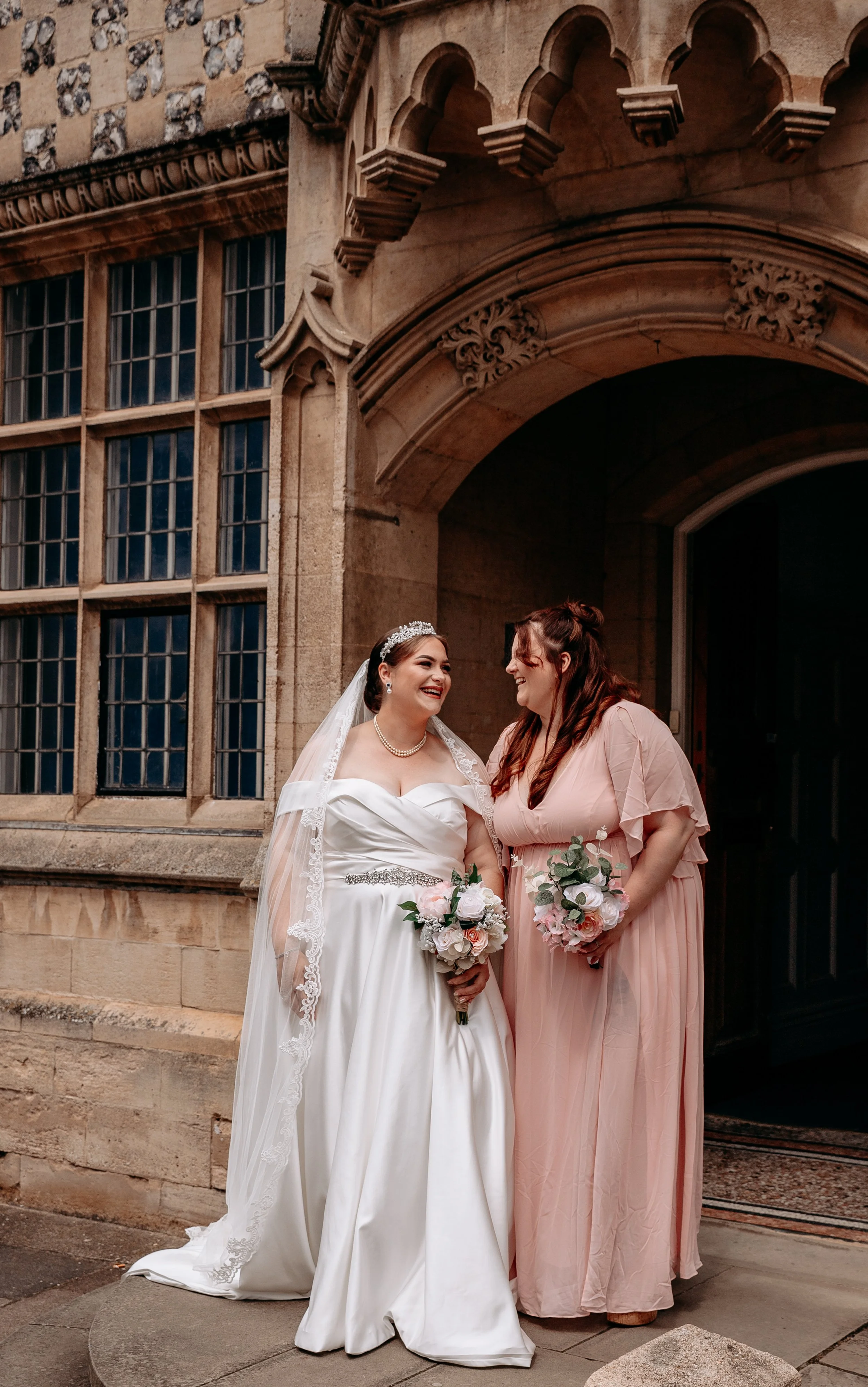 A bride in a white wedding gown and a bridesmaid in a pink dress standing outside a historic stone building, smiling and holding bouquets.