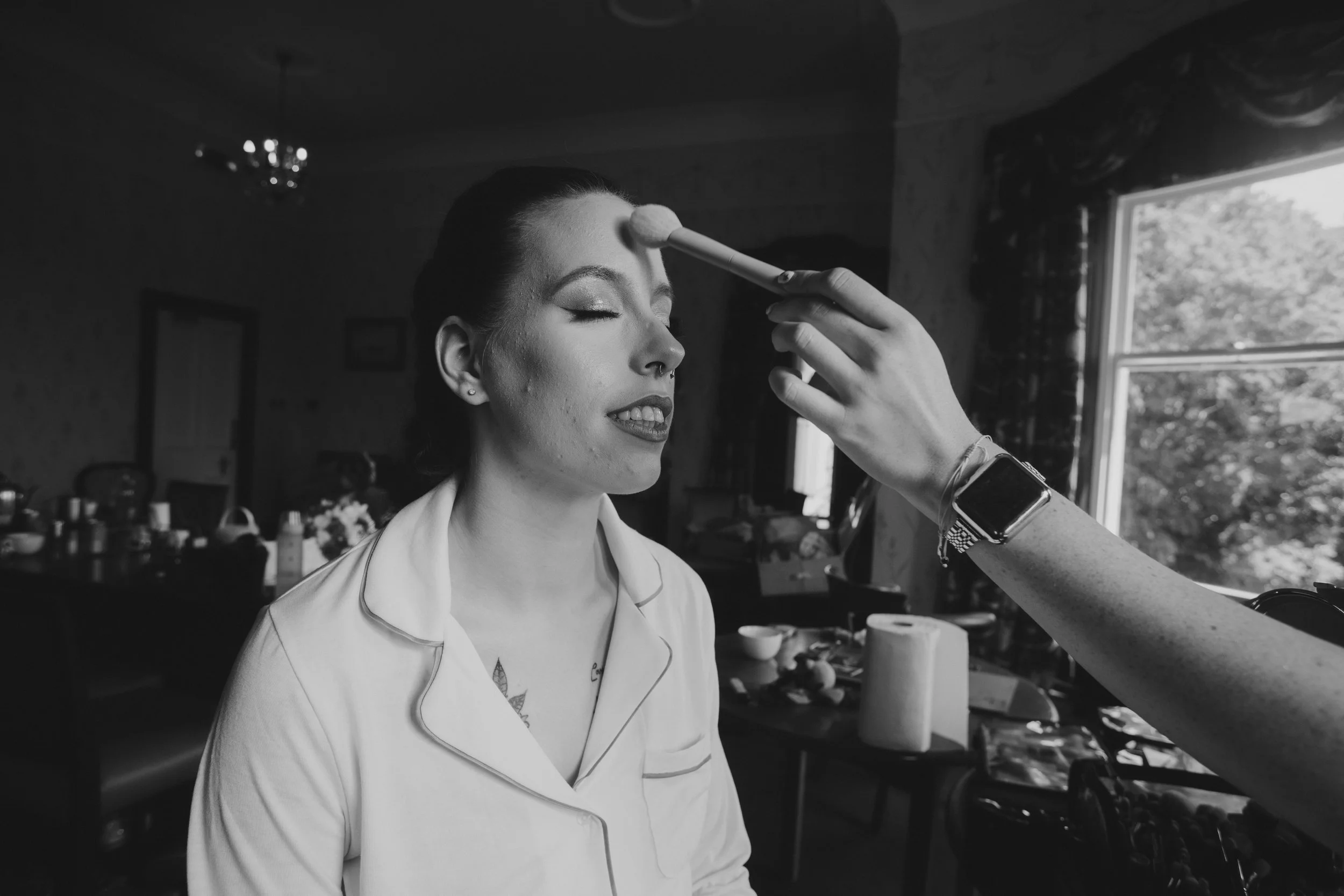 A woman with makeup and a tattoo on her chest receives makeup application in a well-lit room with large windows and curtains.