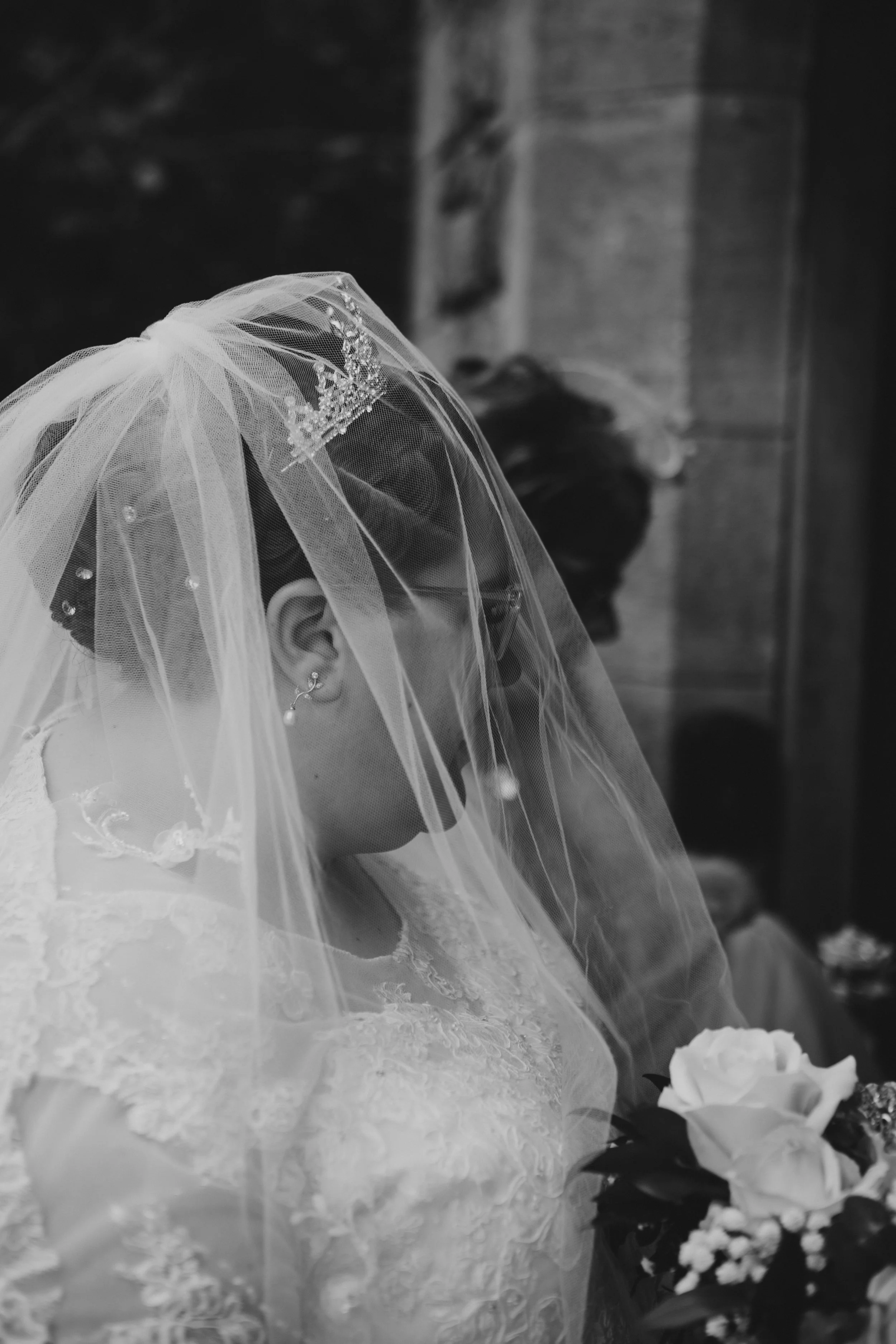 Black and white photo of a bride wearing a lace wedding dress, veil, and tiara, holding a bouquet of flowers, with her head bowed.