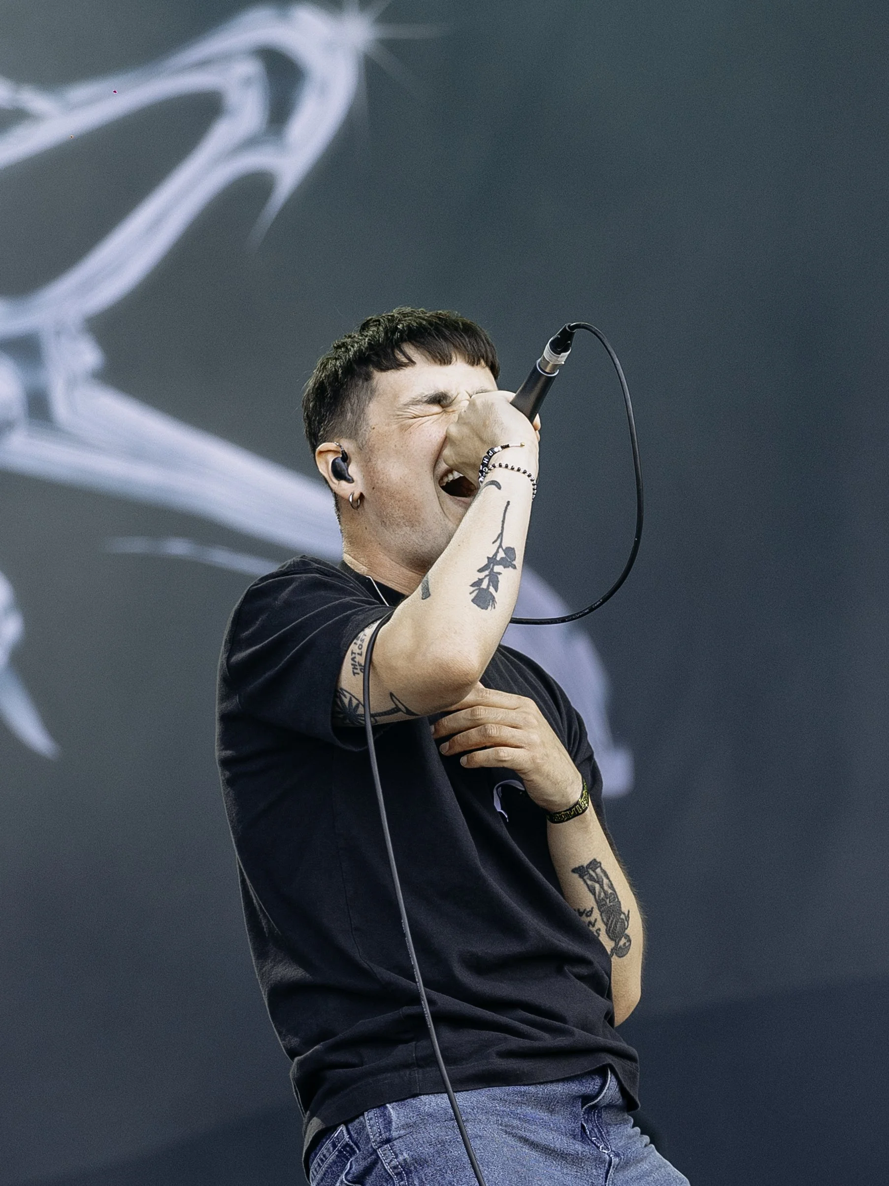 A young man with tattoos singing passionately into a microphone on stage, wearing a black t-shirt and blue jeans.