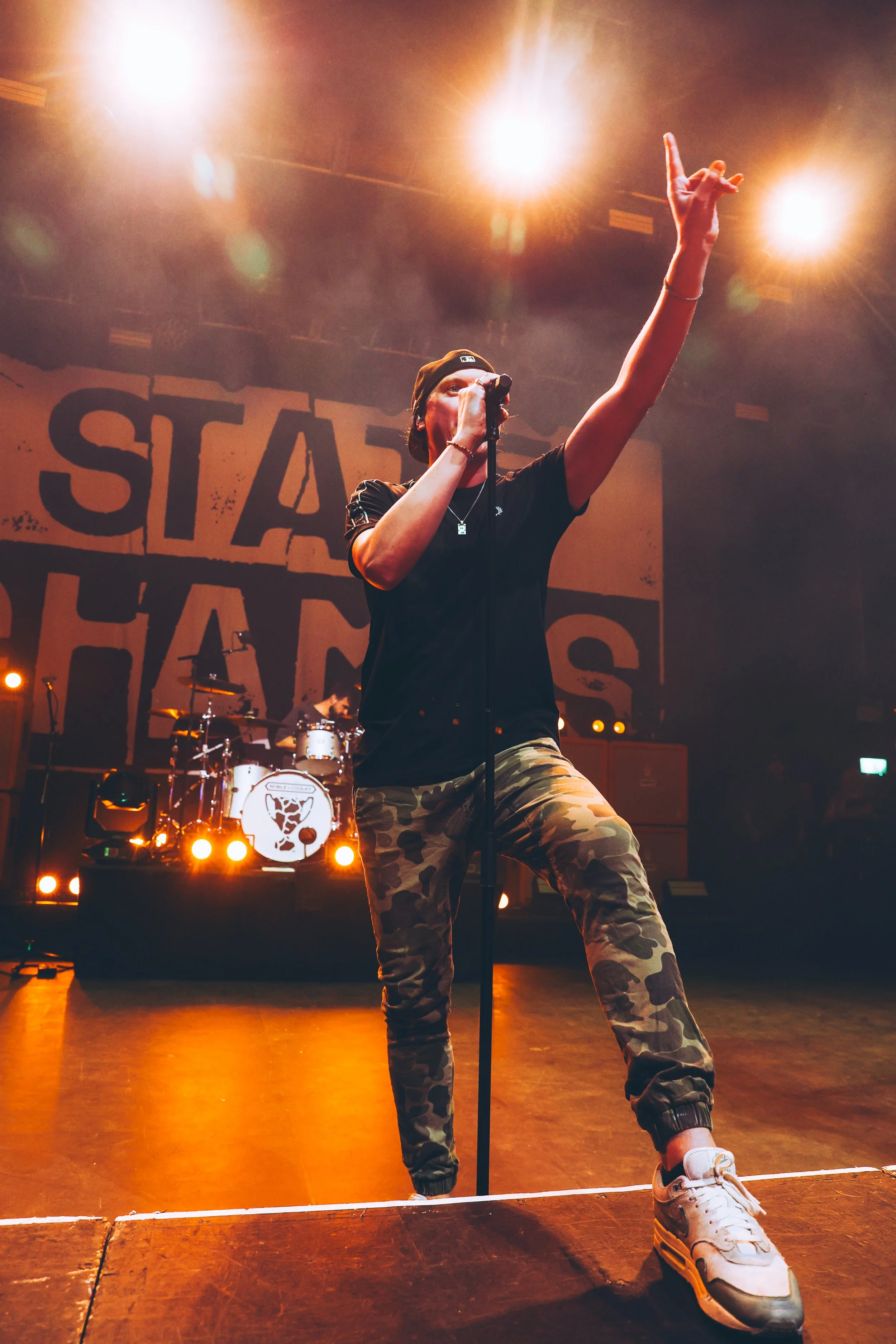 A male singer on stage with camo pants and a black shirt, holding a microphone, raising his right arm, with a band playing drums in the background, stage lights overhead, and a large backdrop with the words 'STAY' and 'HANDS' visible.