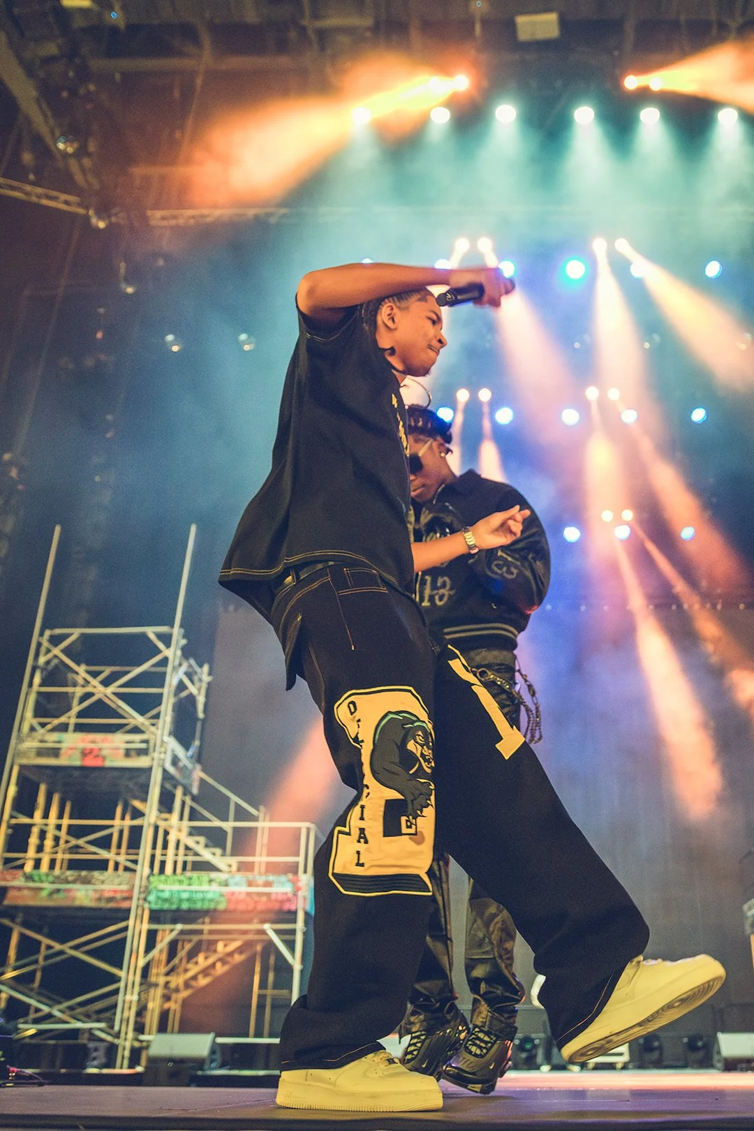 Two performers on stage holding microphones, colorful stage lights, scaffolding in background.