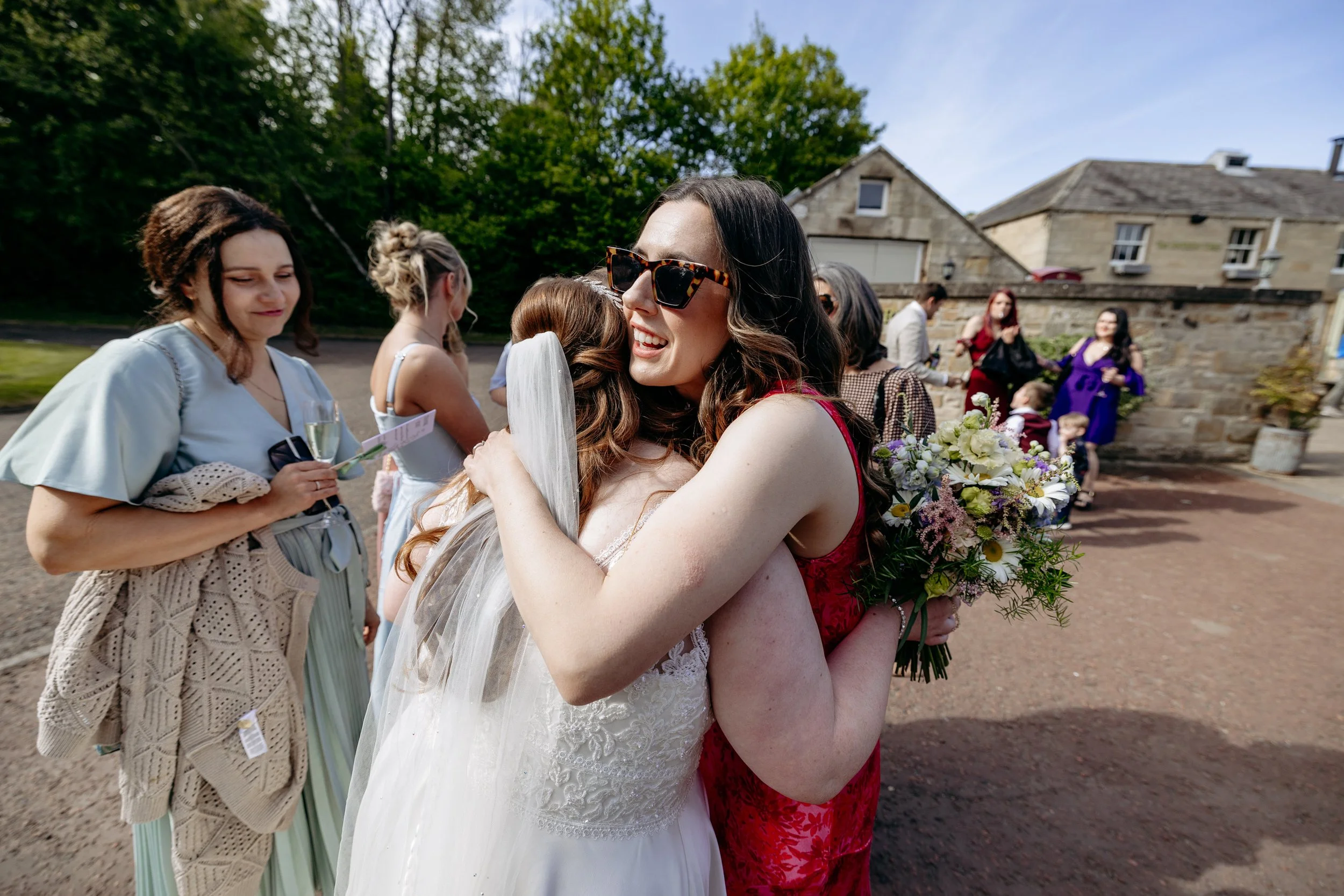 Two women embracing at a wedding, one in a wedding dress and the other in a red dress, holding a bouquet, with other guests in the background outdoors.