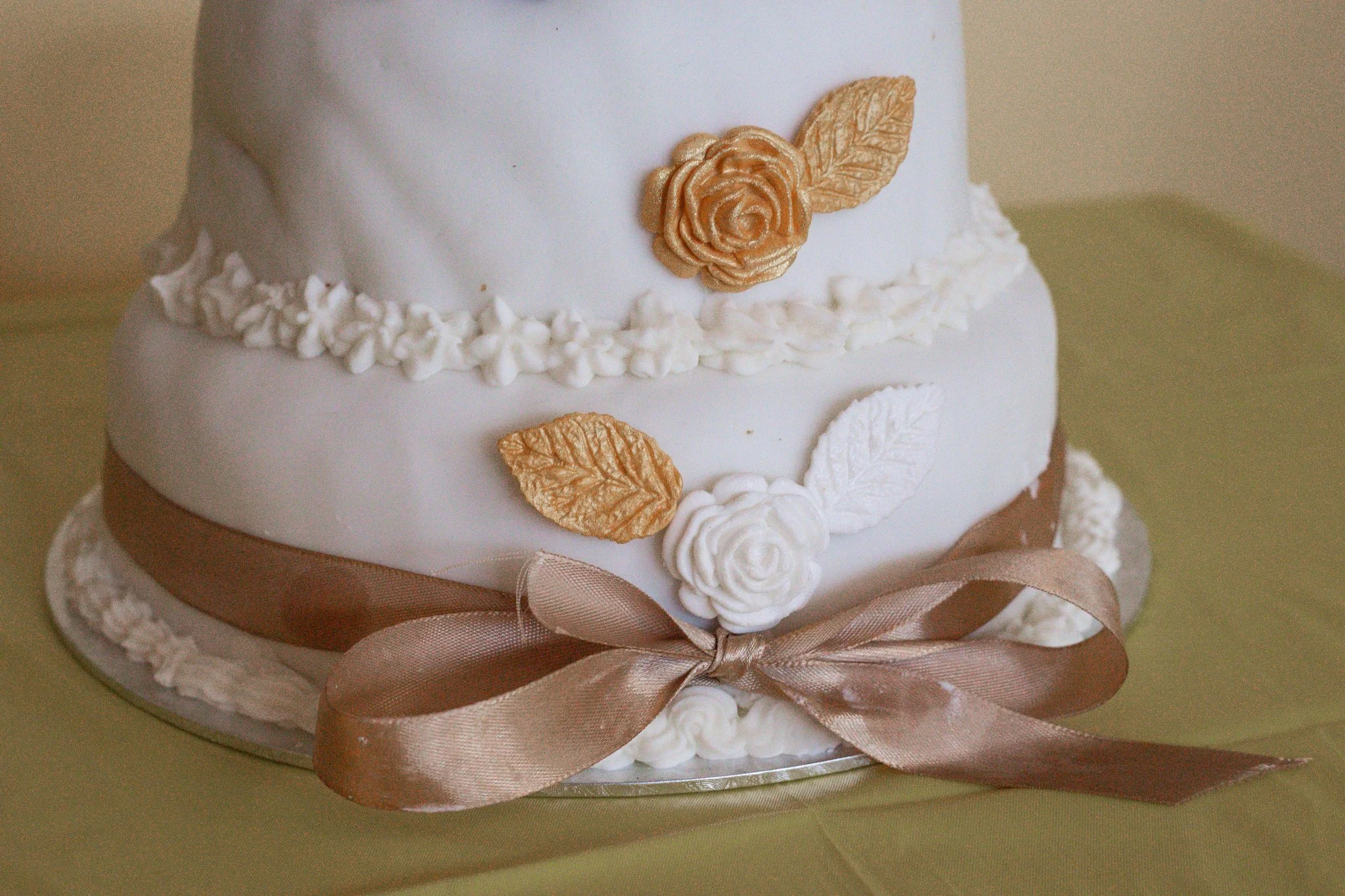 A two-tiered white wedding cake decorated with gold and white edible flowers, gold leaves, and a brown satin ribbon tied in a bow around the bottom tier, sitting on a green tablecloth.
