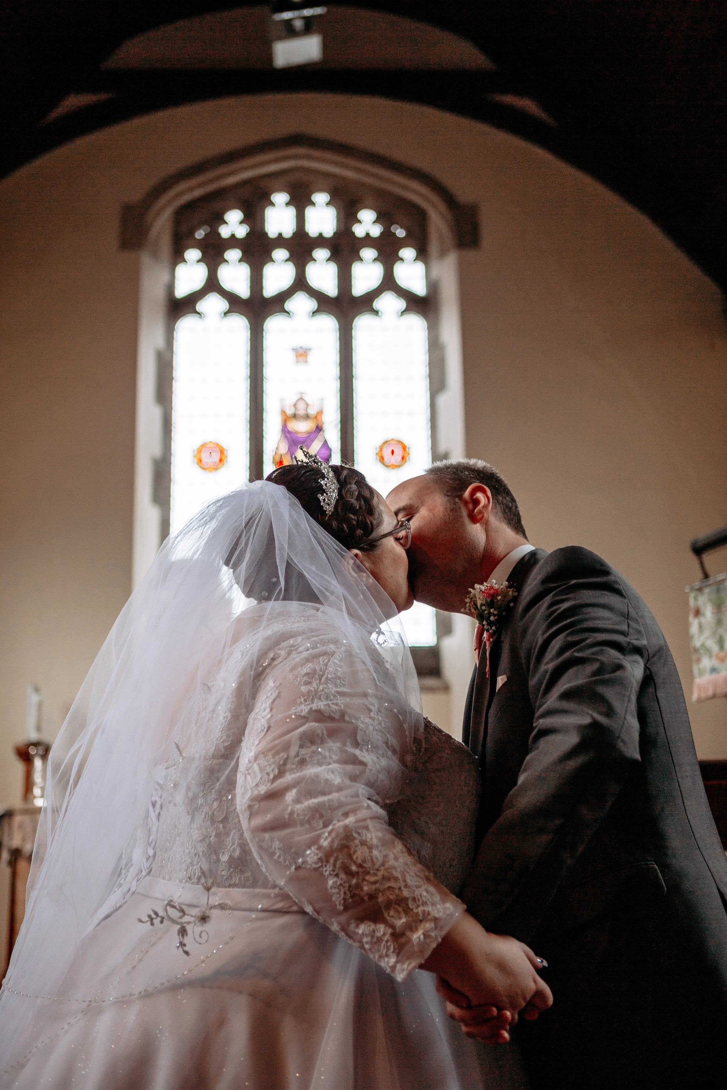 A bride and groom share a kiss during their wedding ceremony inside a church, with a stained glass window behind them.