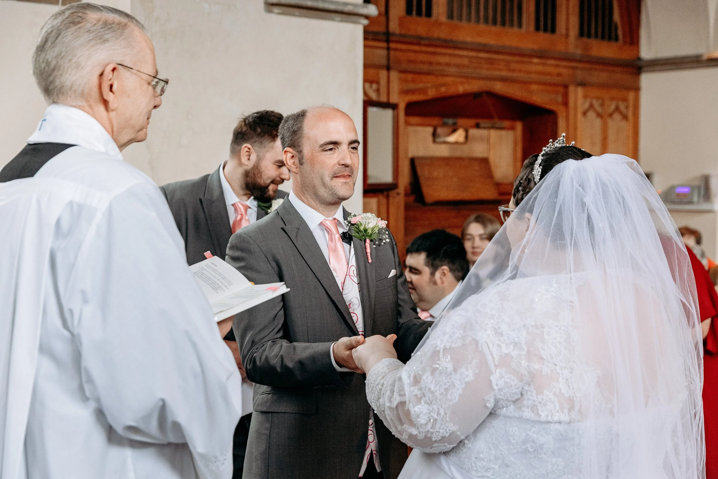 A couple getting married during a church wedding ceremony. The groom is holding hands with the bride, and both are dressed in formal wedding attire. The officiant and guests are visible in the background.