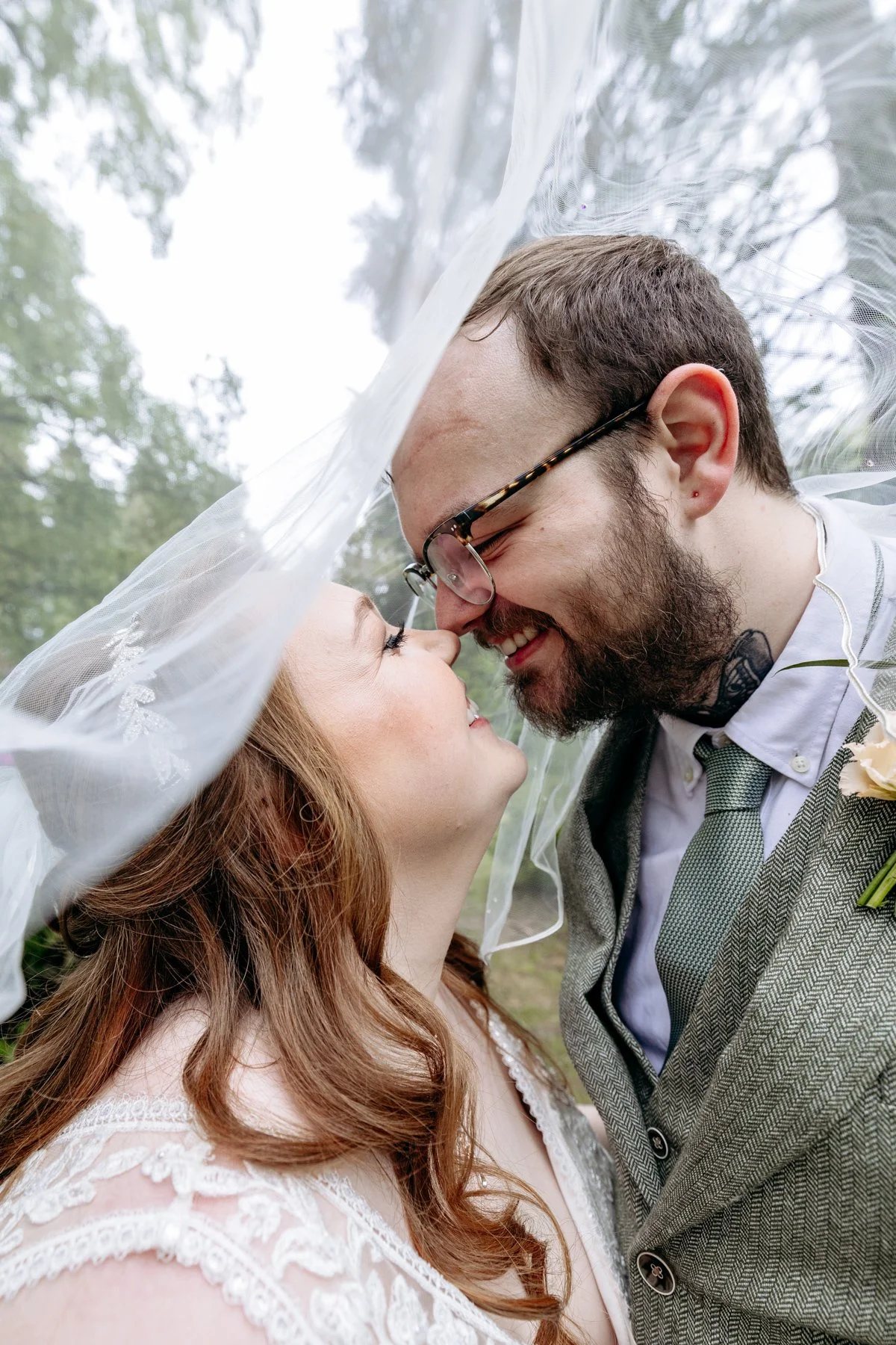 A couple sharing a close, joyful moment under a wedding veil outdoors, surrounded by trees.