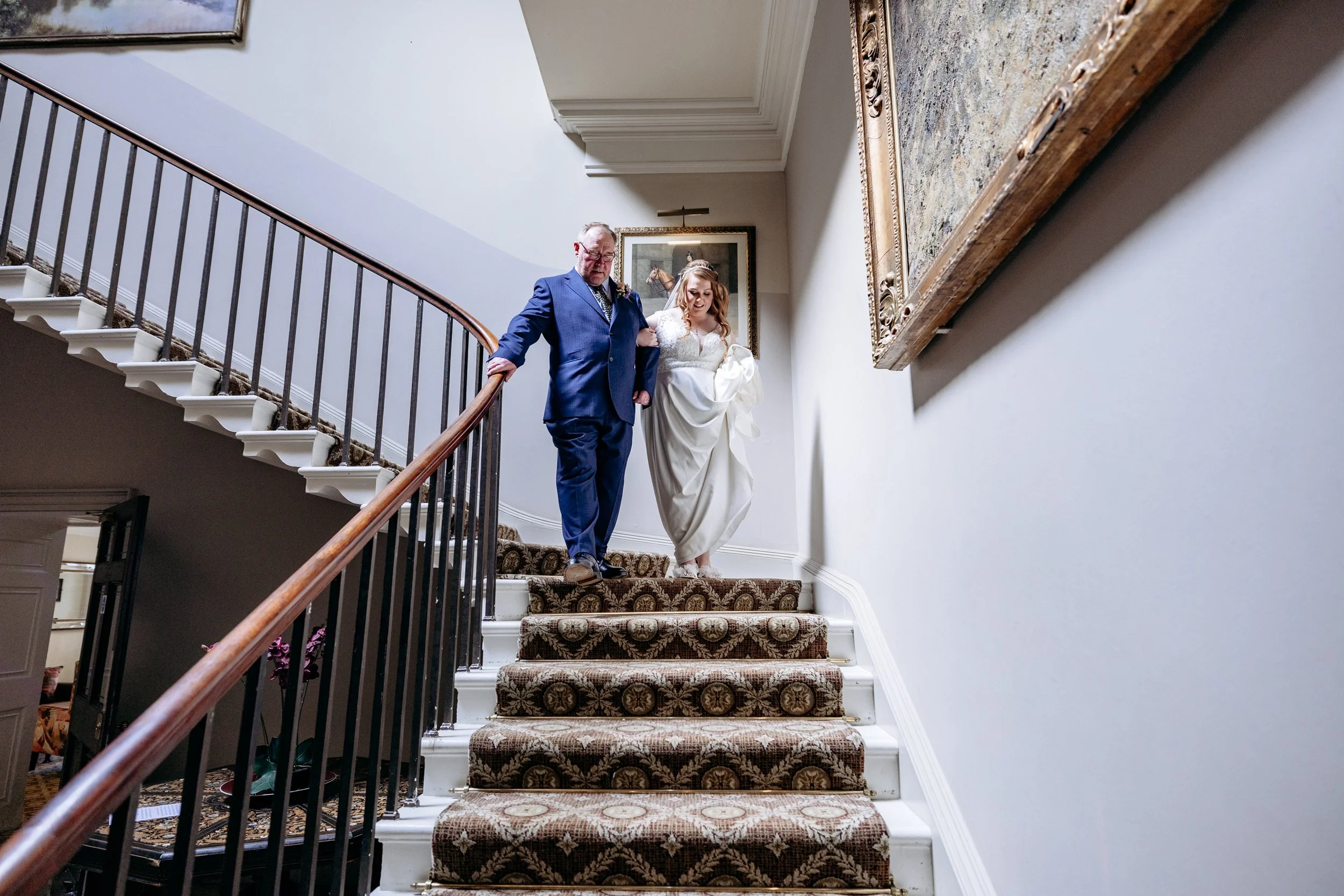 A bride in a white wedding dress being assisted by a man in a blue suit descending a staircase.