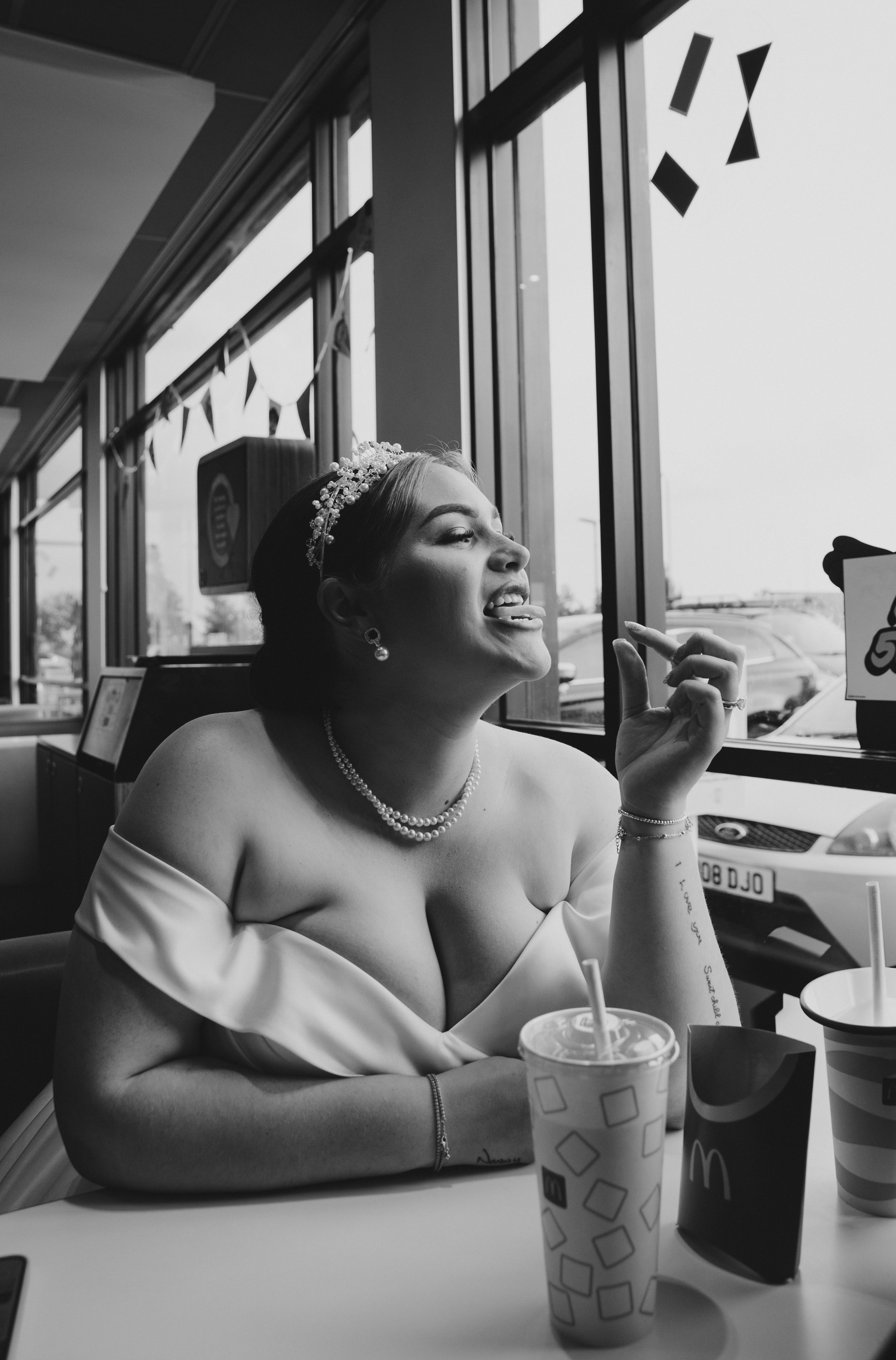Black and white photo of a woman sitting at a fast-food restaurant table, wearing a wedding dress and pearl jewelry, looking out the window with her tongue out, surrounded by McDonald's cups and packaging.