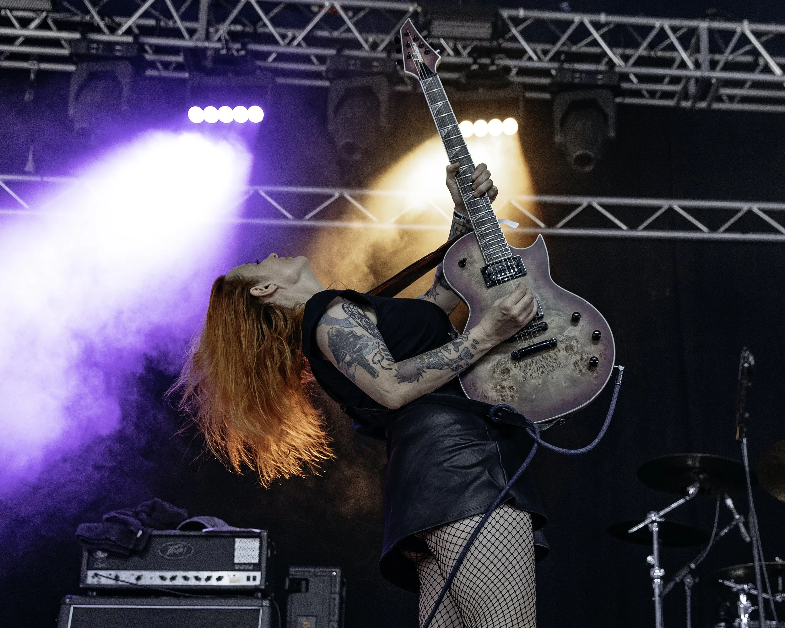 Red-haired female guitarist performing on stage with purple and orange lighting, wearing a black t-shirt, fishnet stockings, and black leather skirt.