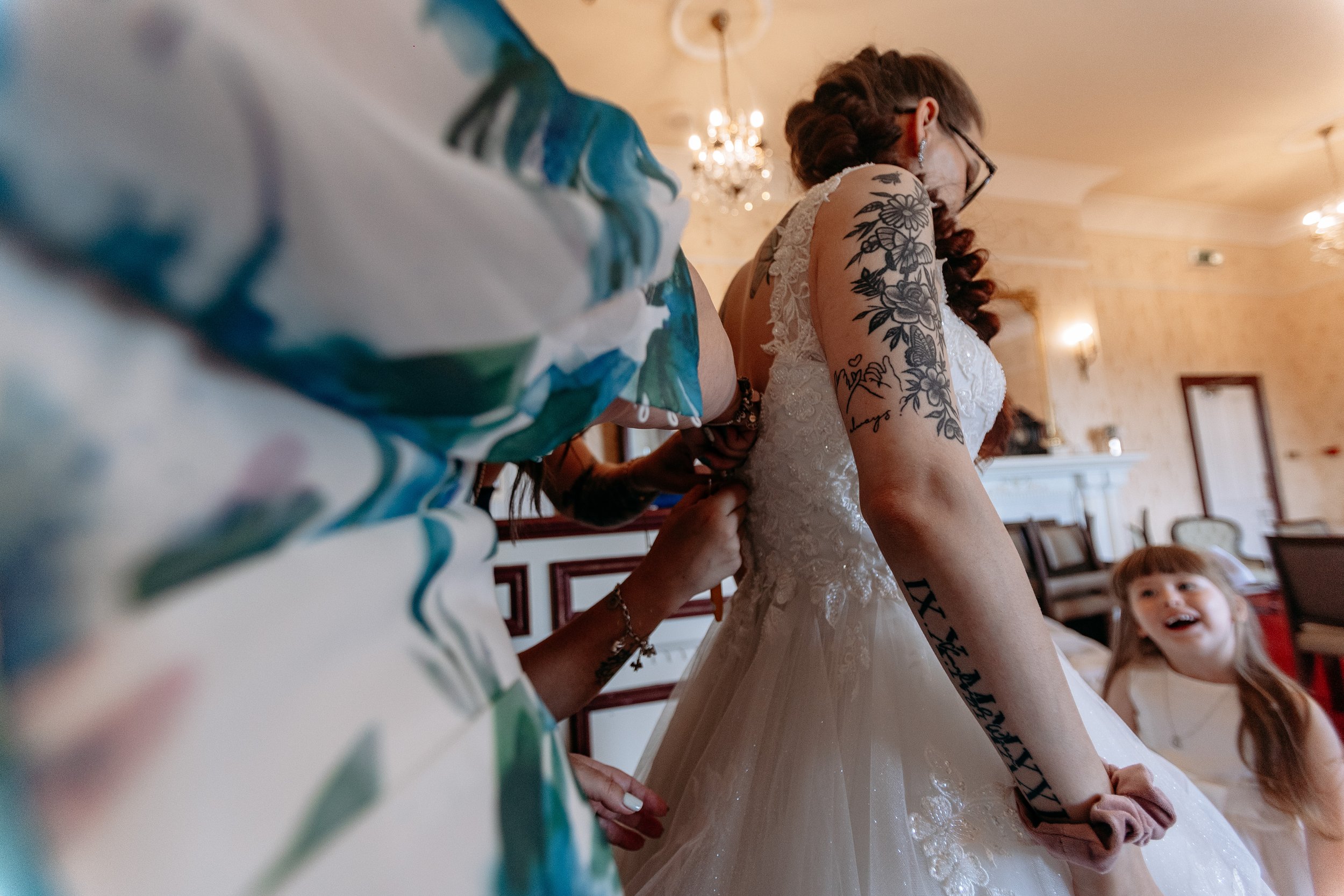 A woman in a wedding dress is being assisted, with a young girl smiling nearby in a decorated indoor setting.