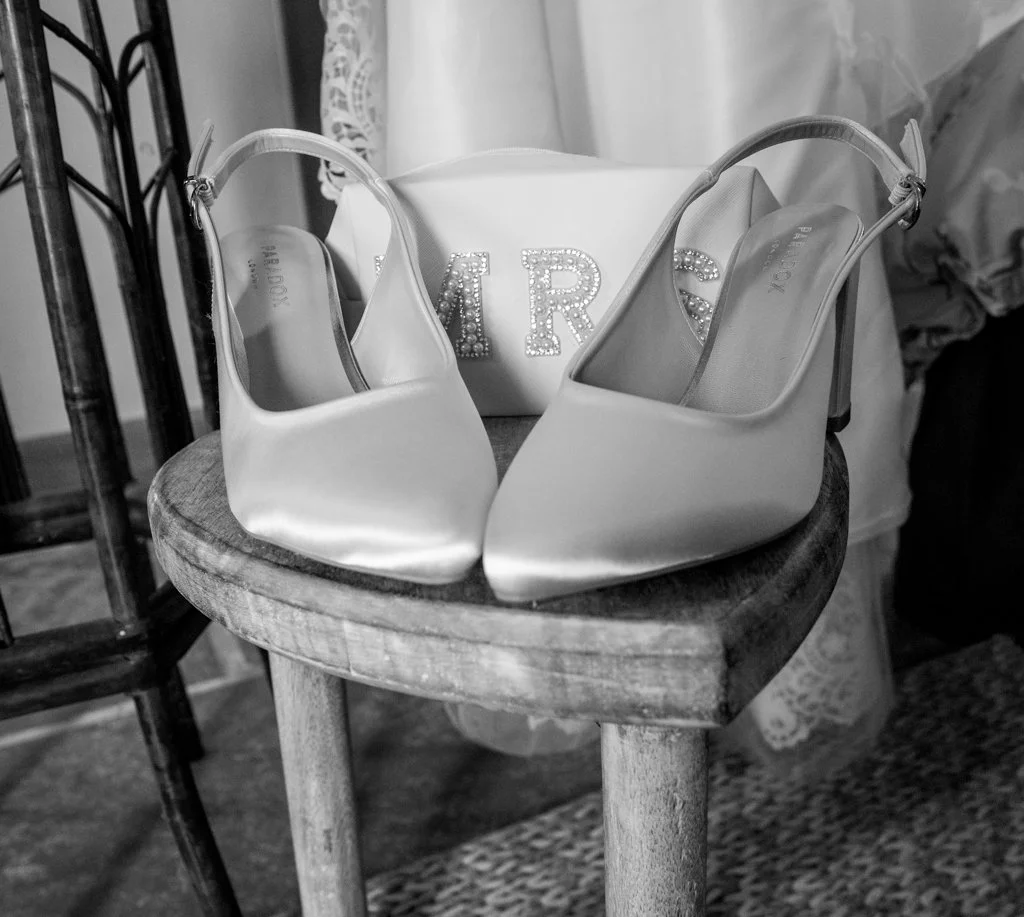 A pair of white high-heeled shoes and a white clutch bag with jewelry spelling 'TRS' on it, placed on a wooden stool.