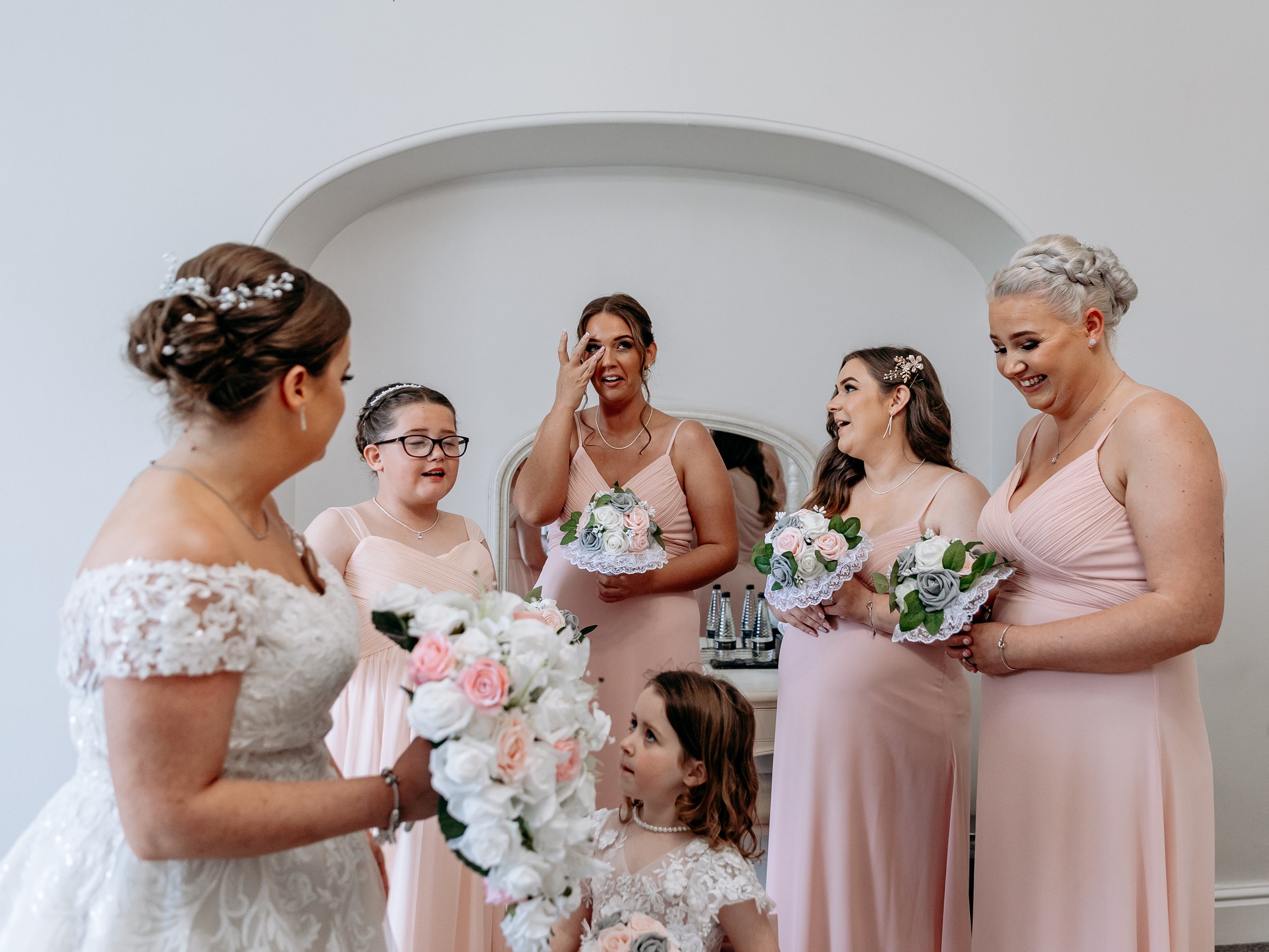 A bride in a white lace wedding gown holding a large bouquet of white and pink roses, talking to five bridesmaids dressed in light pink gowns, holding smaller bouquets of roses, with a young girl in a white lace dress standing nearby. One bridesmaid 