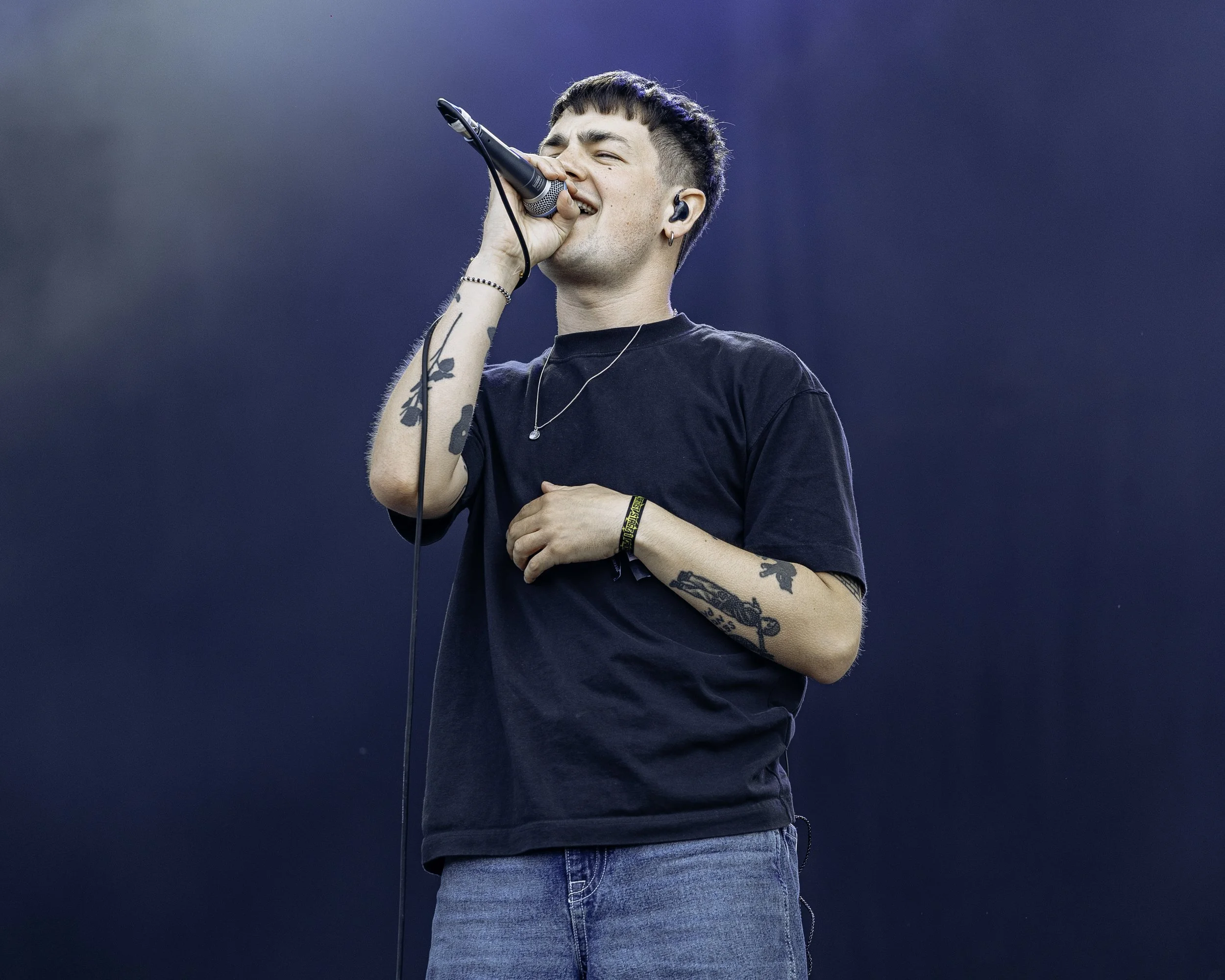 A young man singing into a microphone on stage, wearing a black t-shirt and jeans, with tattoos on his arms and wearing earbuds.