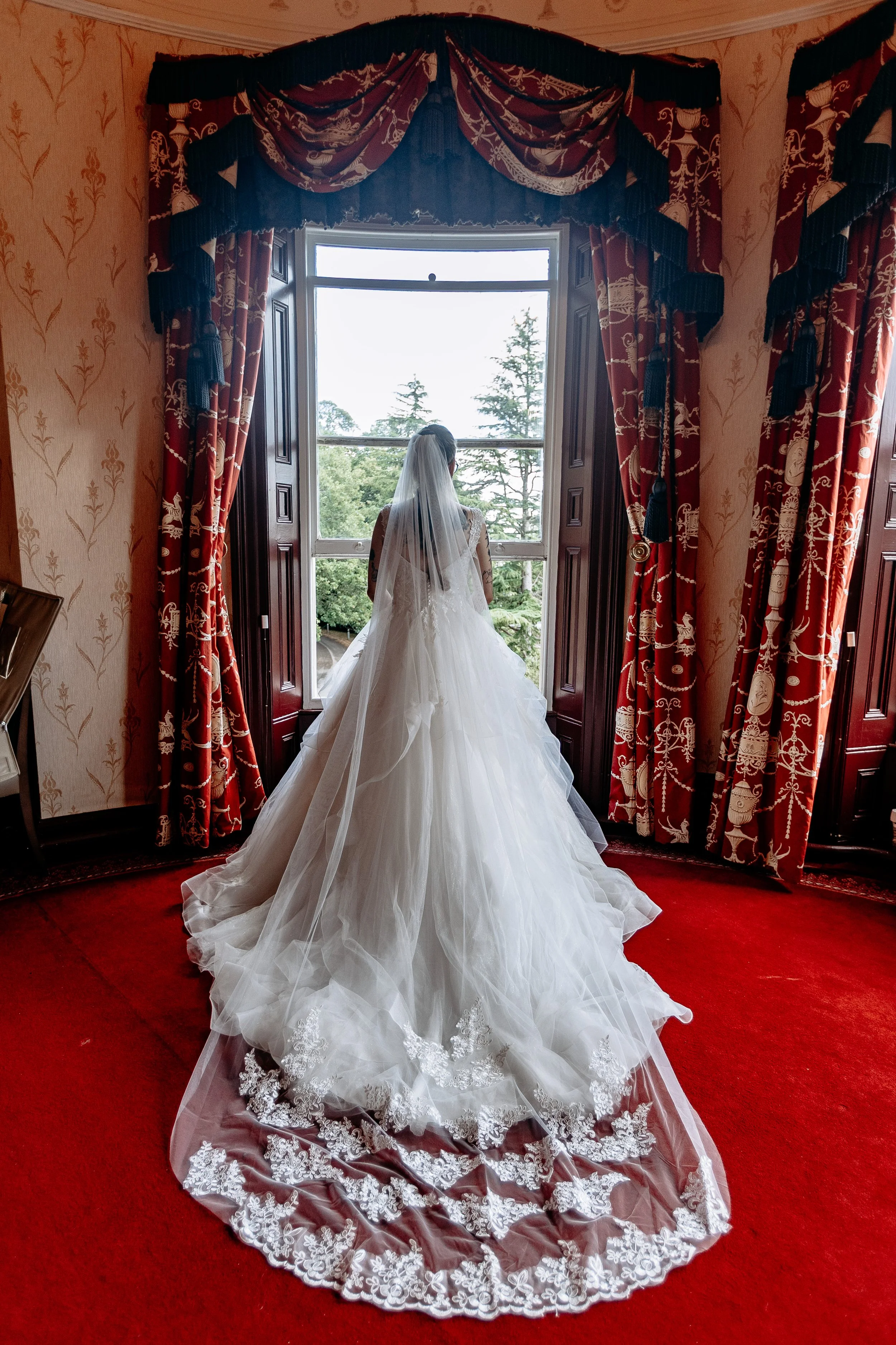A bride in a white wedding gown with a long train and lace details, looking out a large window in a decorated historic room with red carpet, ornate curtains, and patterned wallpaper.
