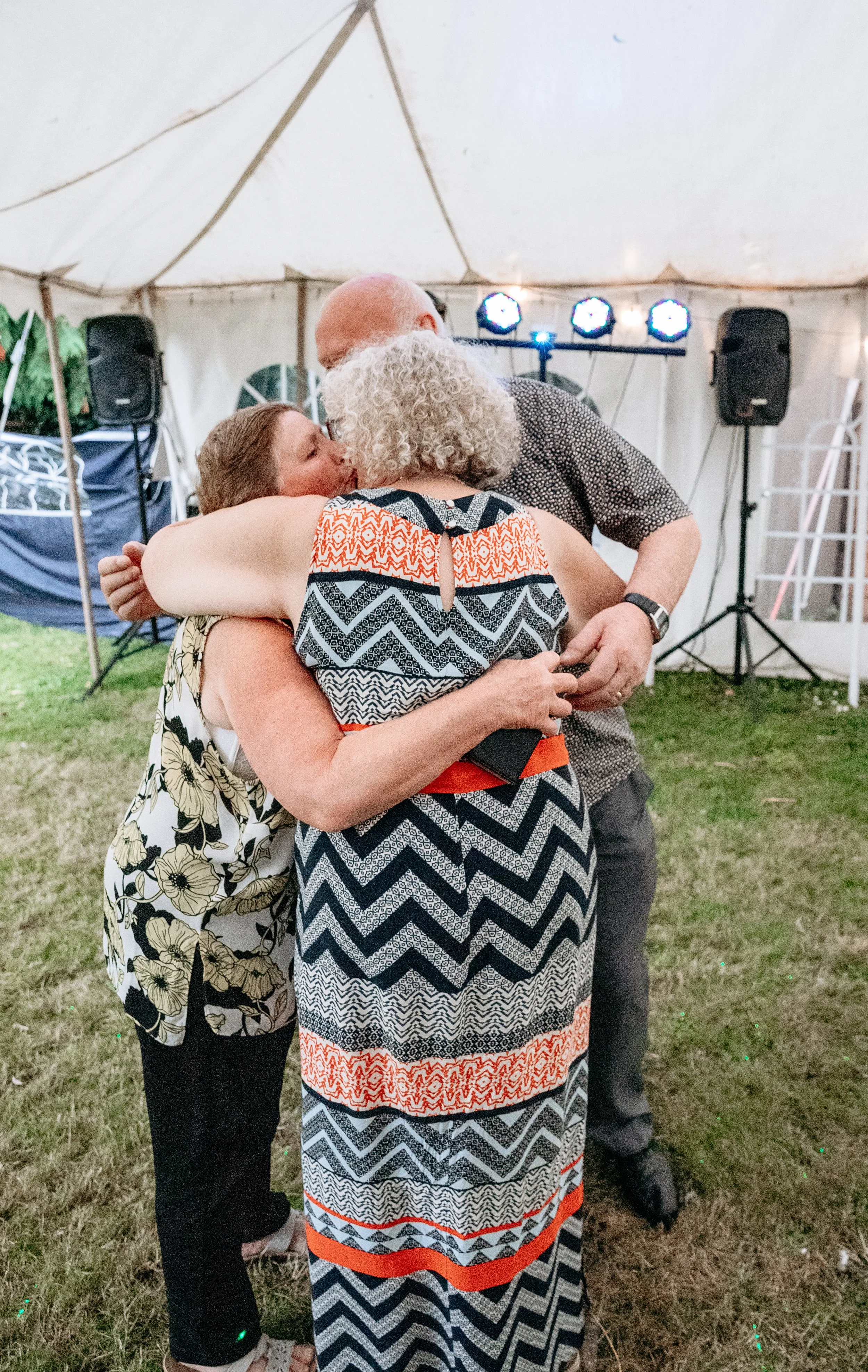 Three people hugging in a tent with lighting and speakers in the background.