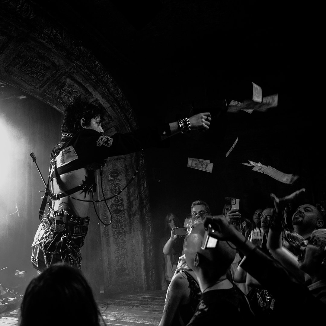 A performer on stage throwing money into a crowd at a concert or nightclub, with audience members taking photos and reaching out.