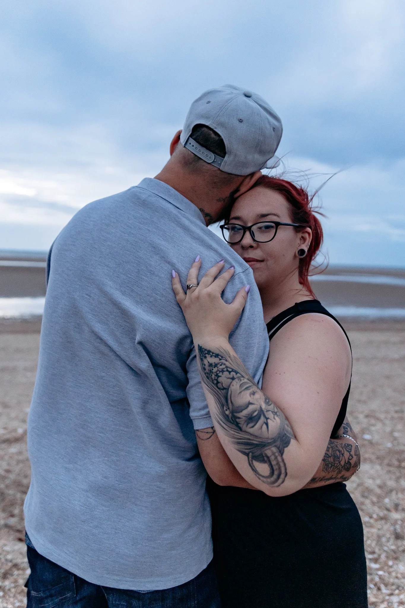 A couple embracing on a beach at sunset, with the man wearing a gray cap and the woman with red hair, glasses, and tattoos on her arm.