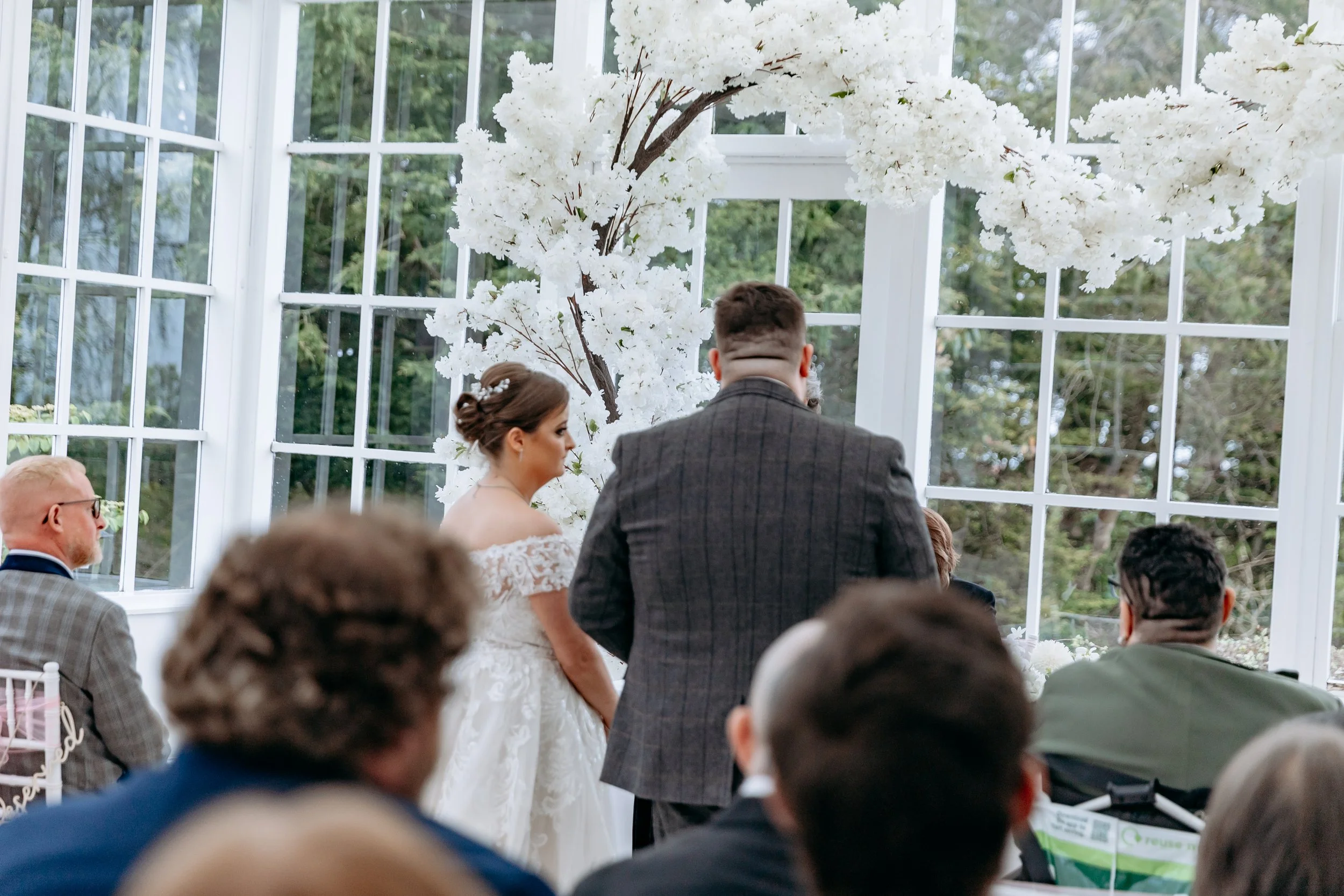 A wedding ceremony taking place in a glass conservatory with white floral decorations and an arbor backdrop, featuring a bride and groom standing before guests inside.