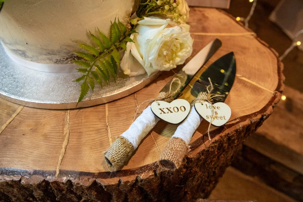 A rustic wooden table with a round log slice top, decorated with a cake, white roses, green fern leaves, and a set of wedding or celebration utensils wrapped in burlap and white cloth, with small wooden tags reading 'XXOO' and 'Love is sweet'.
