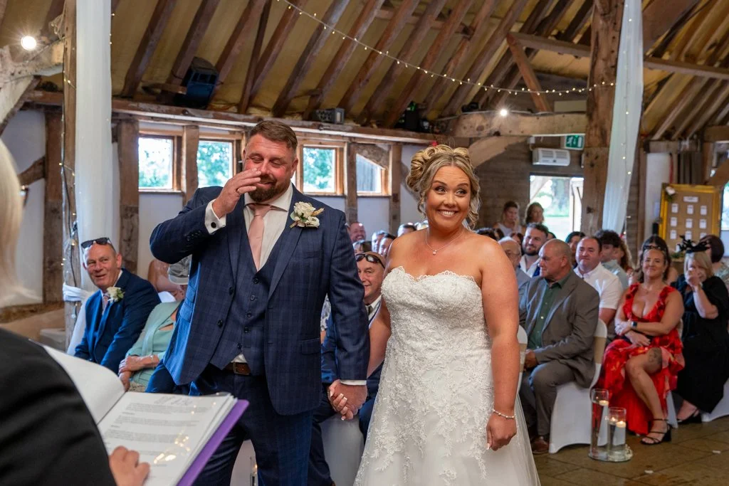 Bride and groom holding hands during their wedding ceremony, surrounded by seated guests in a rustic barn with string lights overhead.