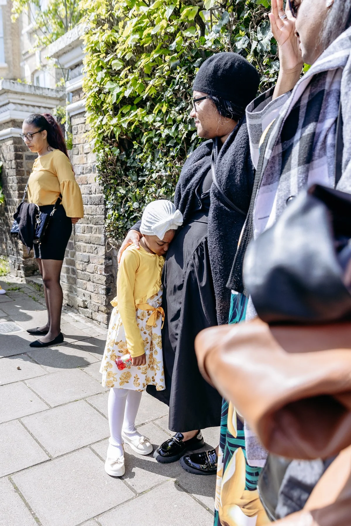 A young girl in a yellow top and floral skirt is hugging an older woman in black, who is wearing glasses and a head covering, while standing on a sidewalk on a sunny day. Other people are nearby, some looking at their phones and standing against a br
