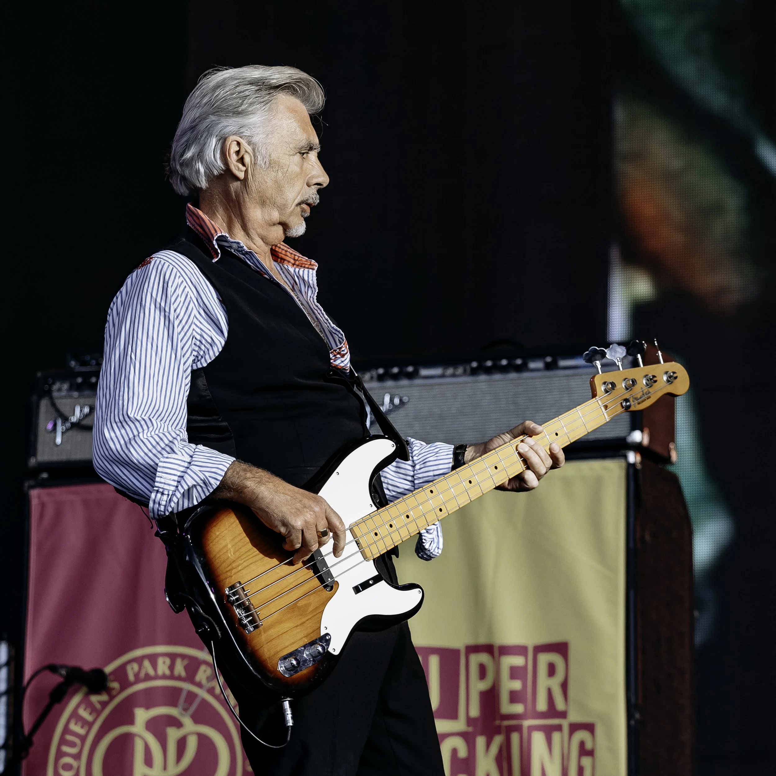 An elderly man with gray hair, wearing a striped shirt and black vest, playing an electric bass guitar on stage.