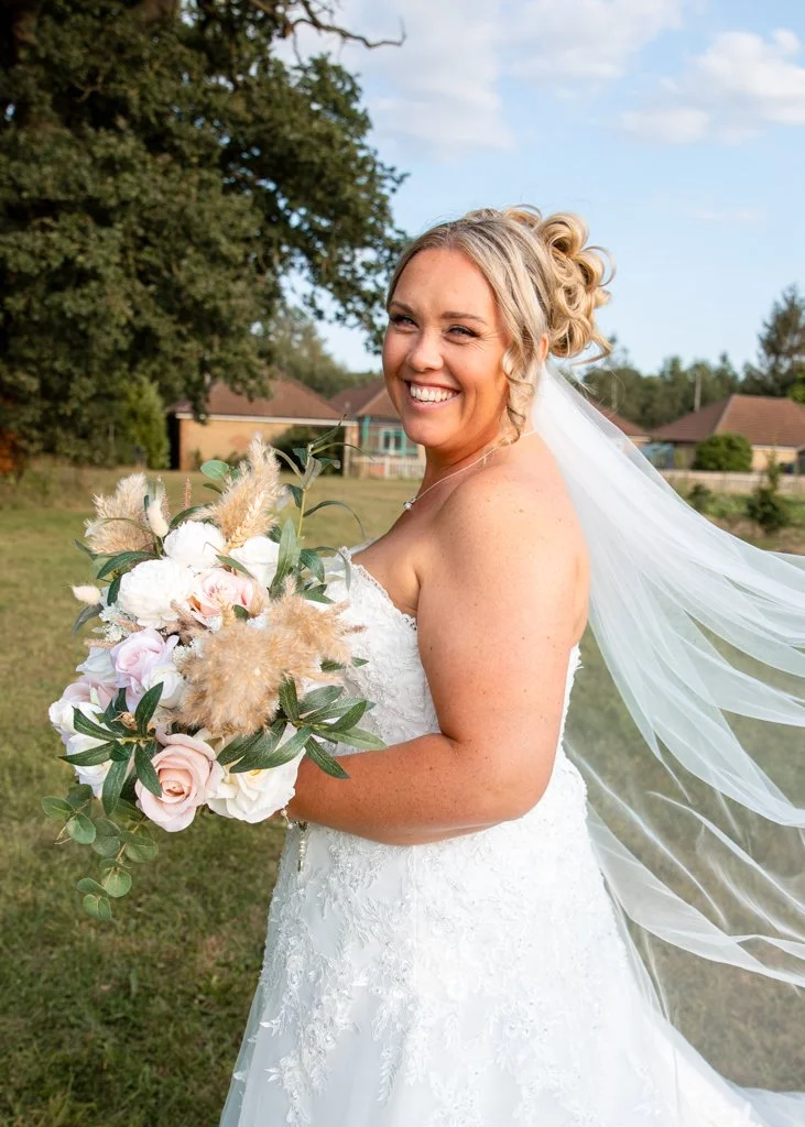 A smiling bride in a wedding dress holding a bouquet of pink and white roses with greenery and beige pampas grass in an outdoor setting.