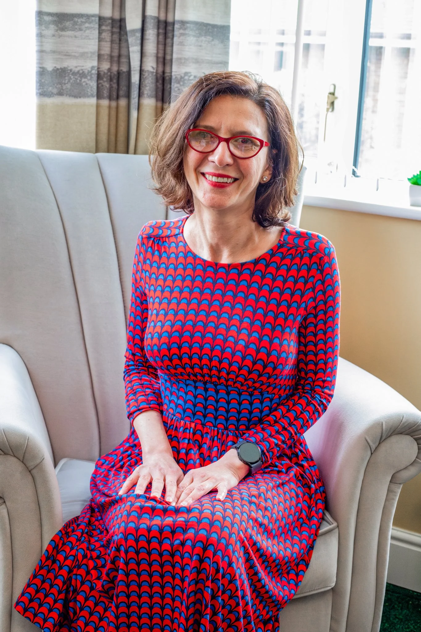 A woman sitting on a beige armchair near a window, wearing a colorful patterned dress and red glasses, smiling at the camera.