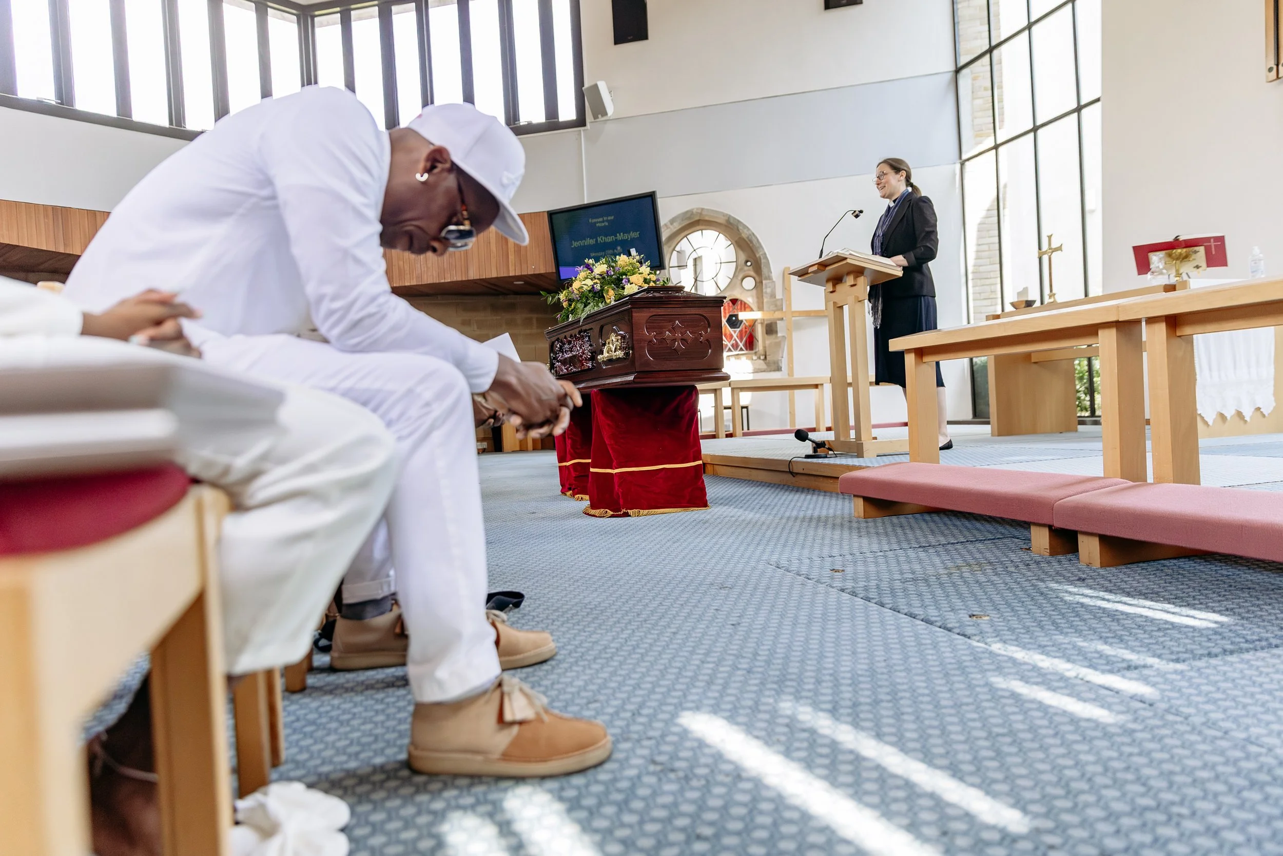A person dressed in white sitting in a church, bowing his head in prayer or reflection, with a woman standing at a pulpit in the background.