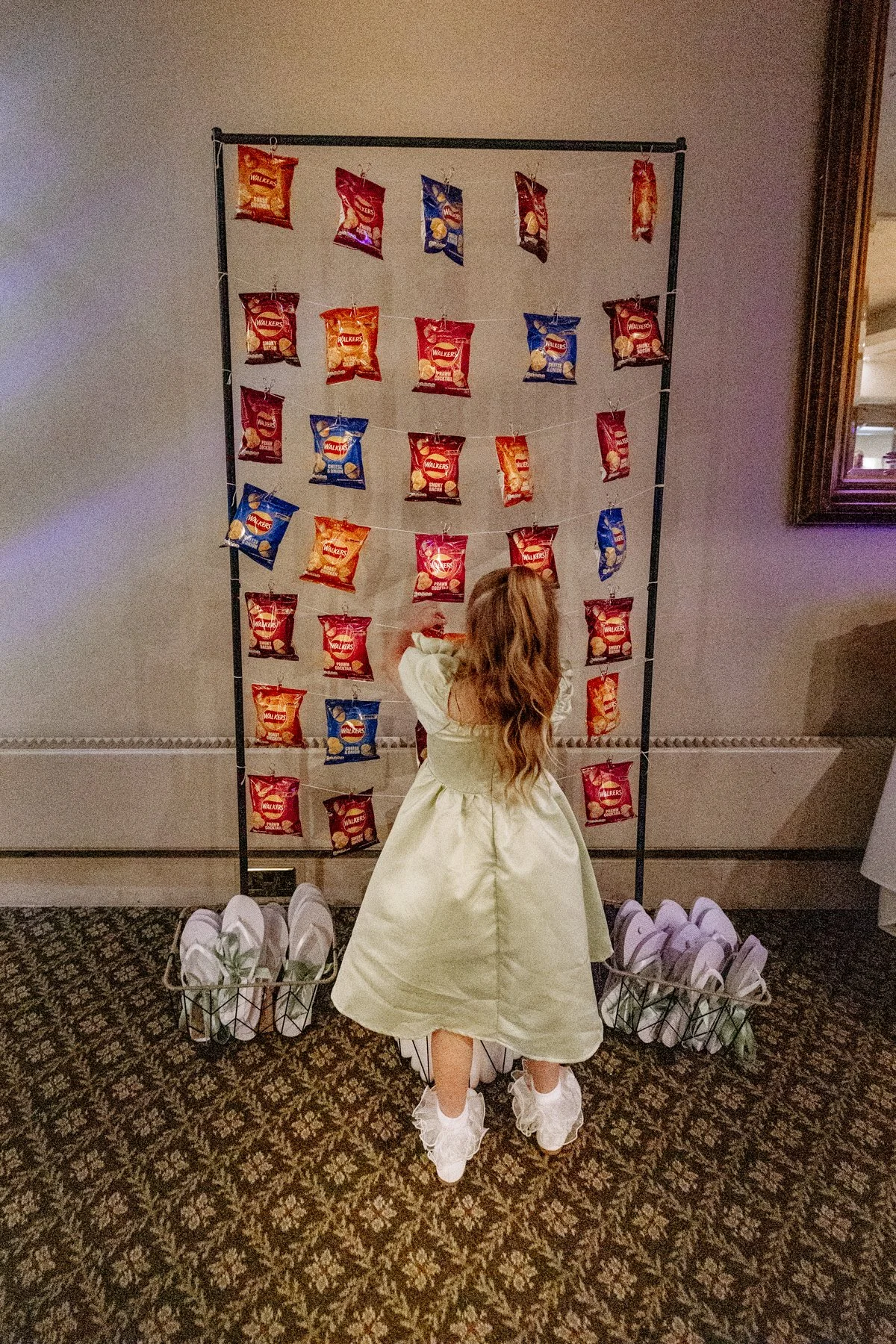 A girl with long hair in a white dress and ruffled shoes is facing away, standing in front of a display of snack bags hanging on a frame. The snack bags are predominantly red, with some blue and orange, and are arranged in rows. The display is set up