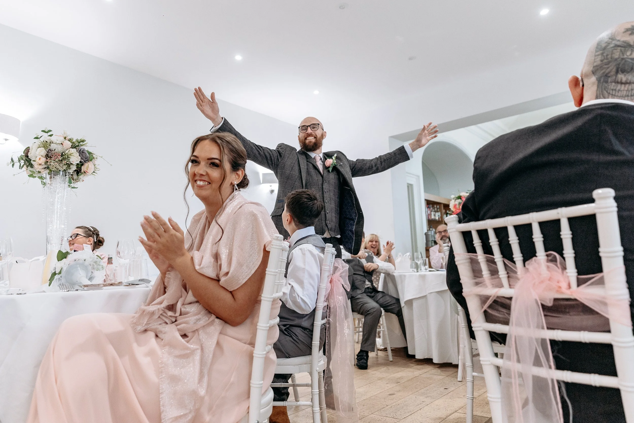 A joyful wedding reception with guests clapping and smiling, including a woman in a pink dress, a man in a gray tuxedo with arms outstretched, and other seated guests at decorated tables.